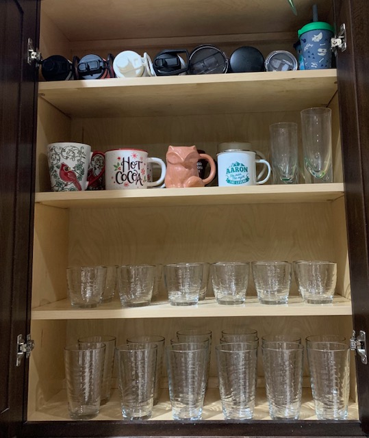 Open cabinet with cups, glasses, and a container on three shelves. Top shelf has small items, middle shelf has mugs, bottom shelves have glasses.