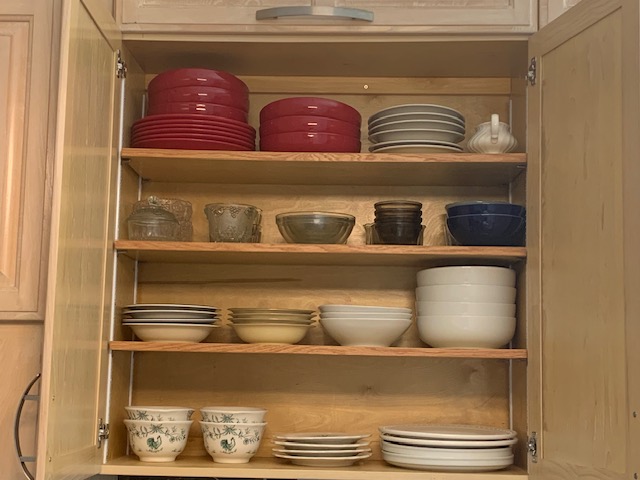 Open wooden cabinet with shelves holding red, gray, black, and blue bowls, plates, and glassware, arranged neatly.