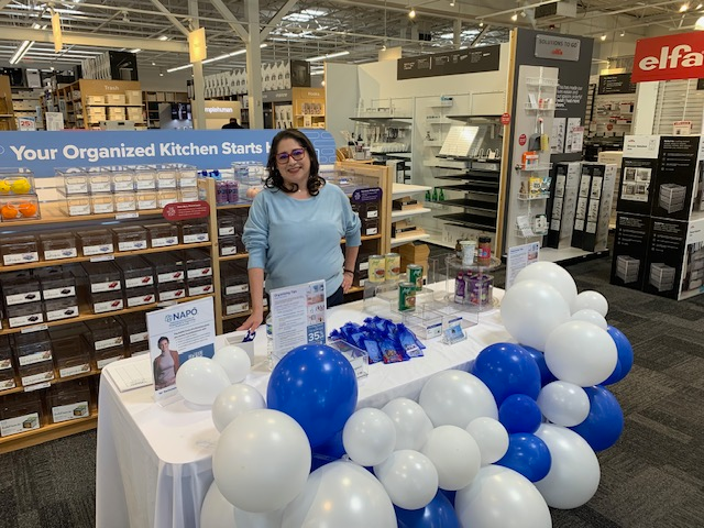 Woman standing behind a table decorated with blue and white balloons in a store, with shelves and products in the background.