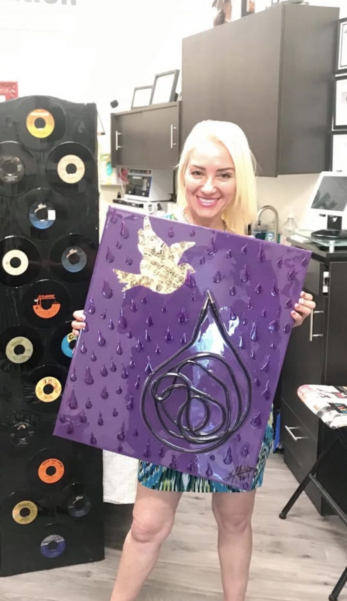Woman with blonde hair holding a purple painting with a crown and abstract design, smiling in a room with speakers and equipment.