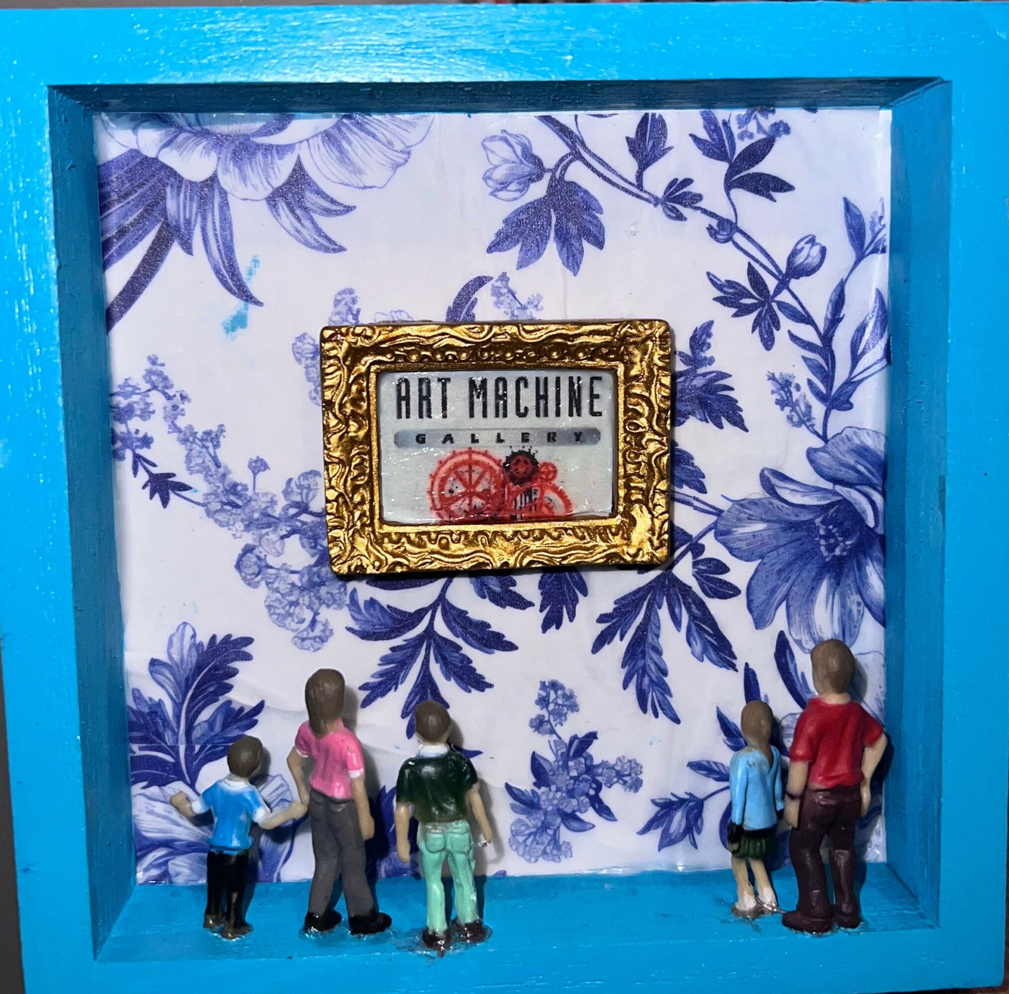 Five small figurines stand in front of a blue and white floral background with a framed sign reading 'Art Machine'.