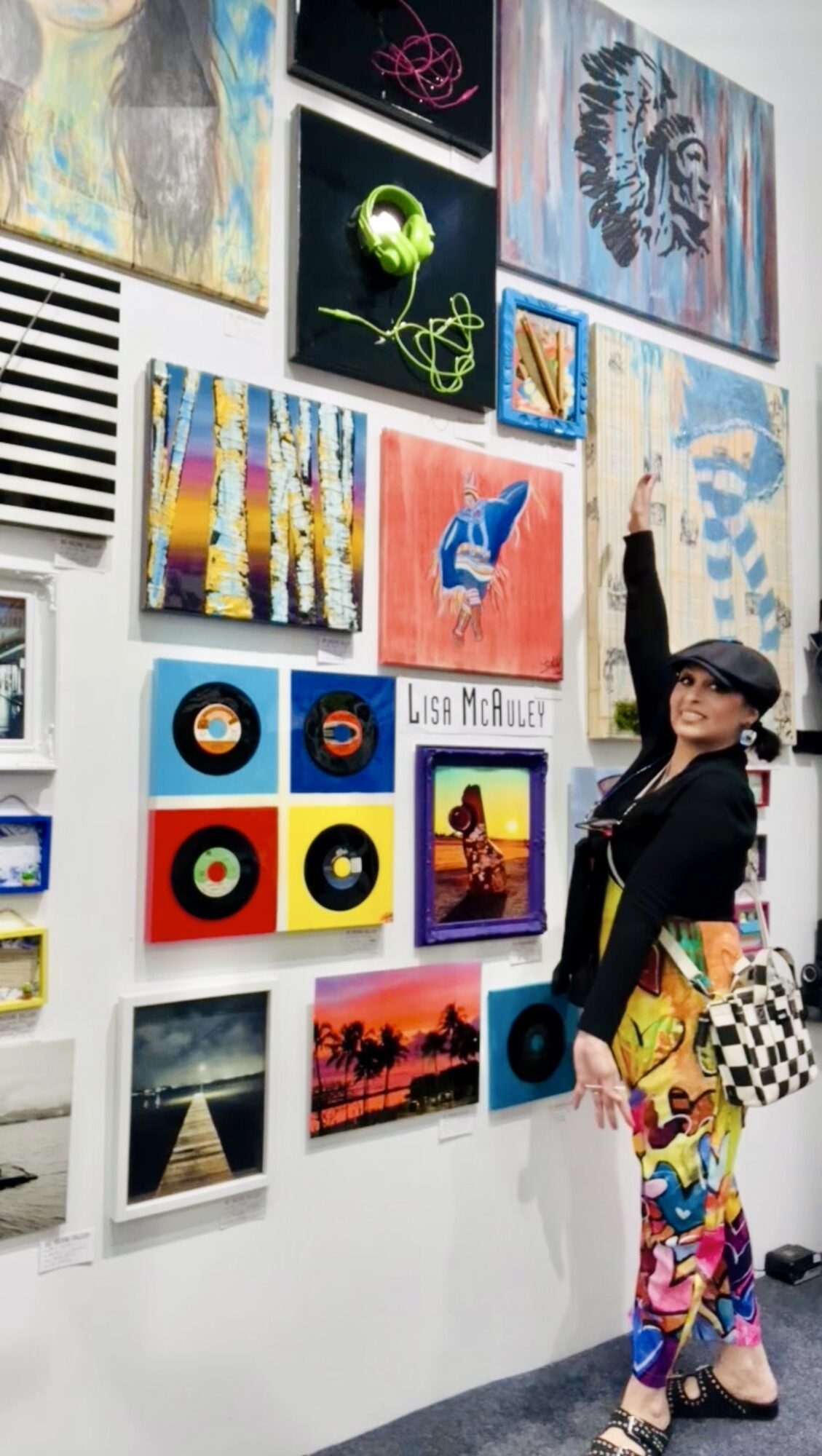 Woman in black top and colorful pants points at artwork on gallery wall, holding a checkered bag.