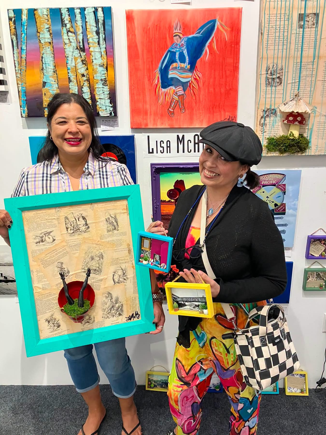 Two women smiling in an art gallery, holding colorful framed artworks, with paintings on the wall behind them.