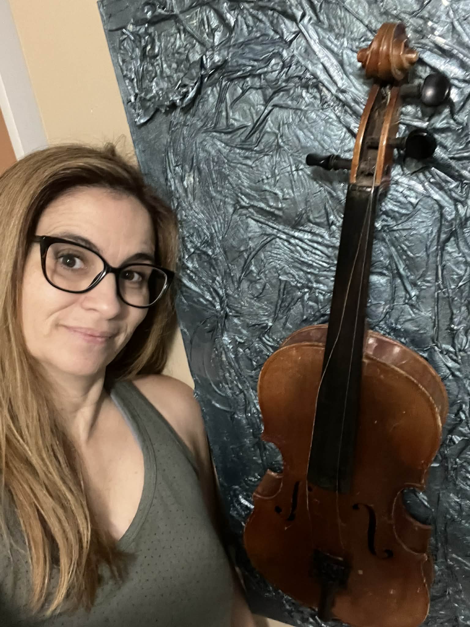 Woman with glasses and long hair taking a selfie next to a large, textured, metallic-looking backdrop and a violin.