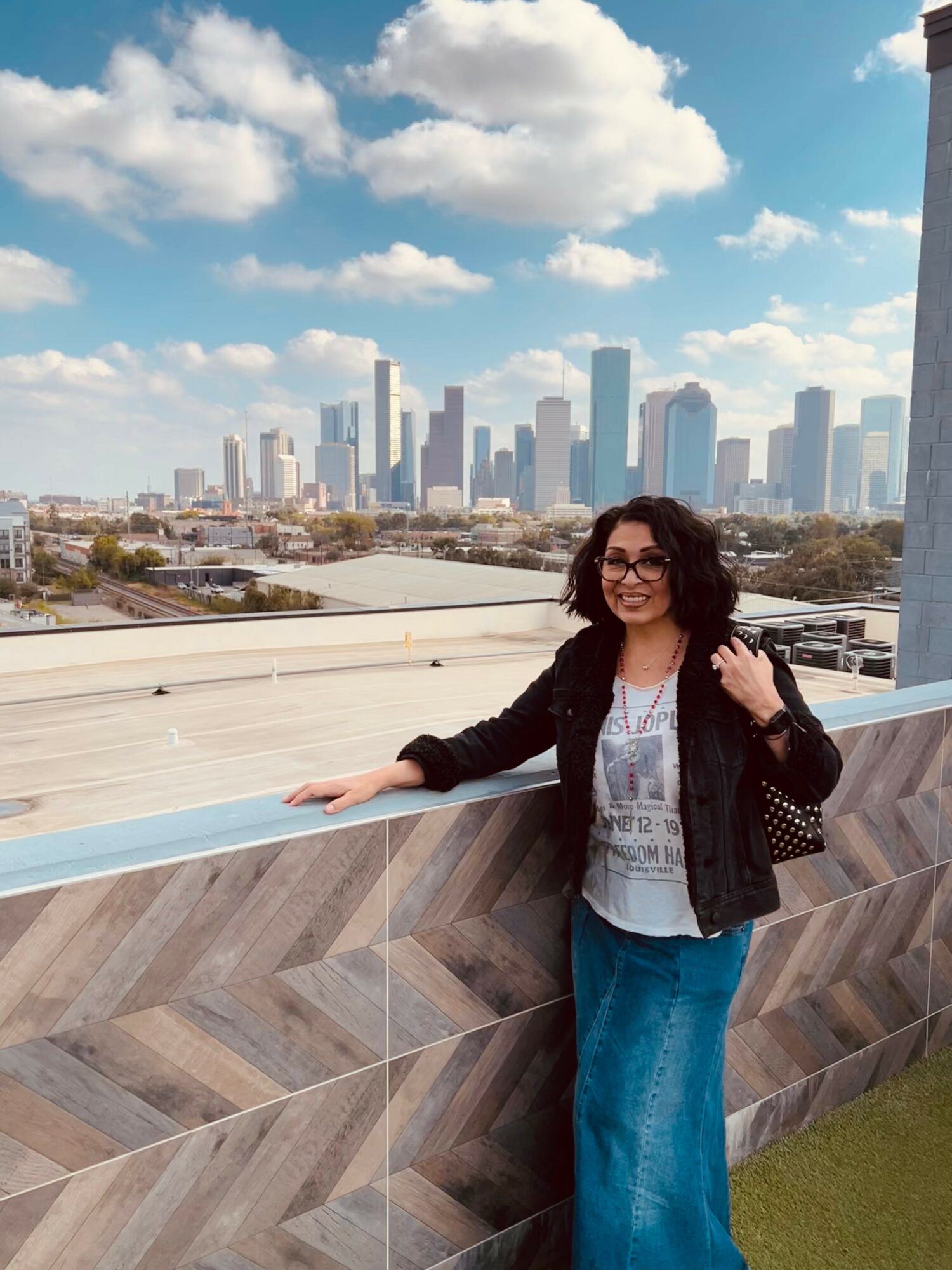 Woman with curly hair and glasses standing outdoors with city skyline and blue sky in background.