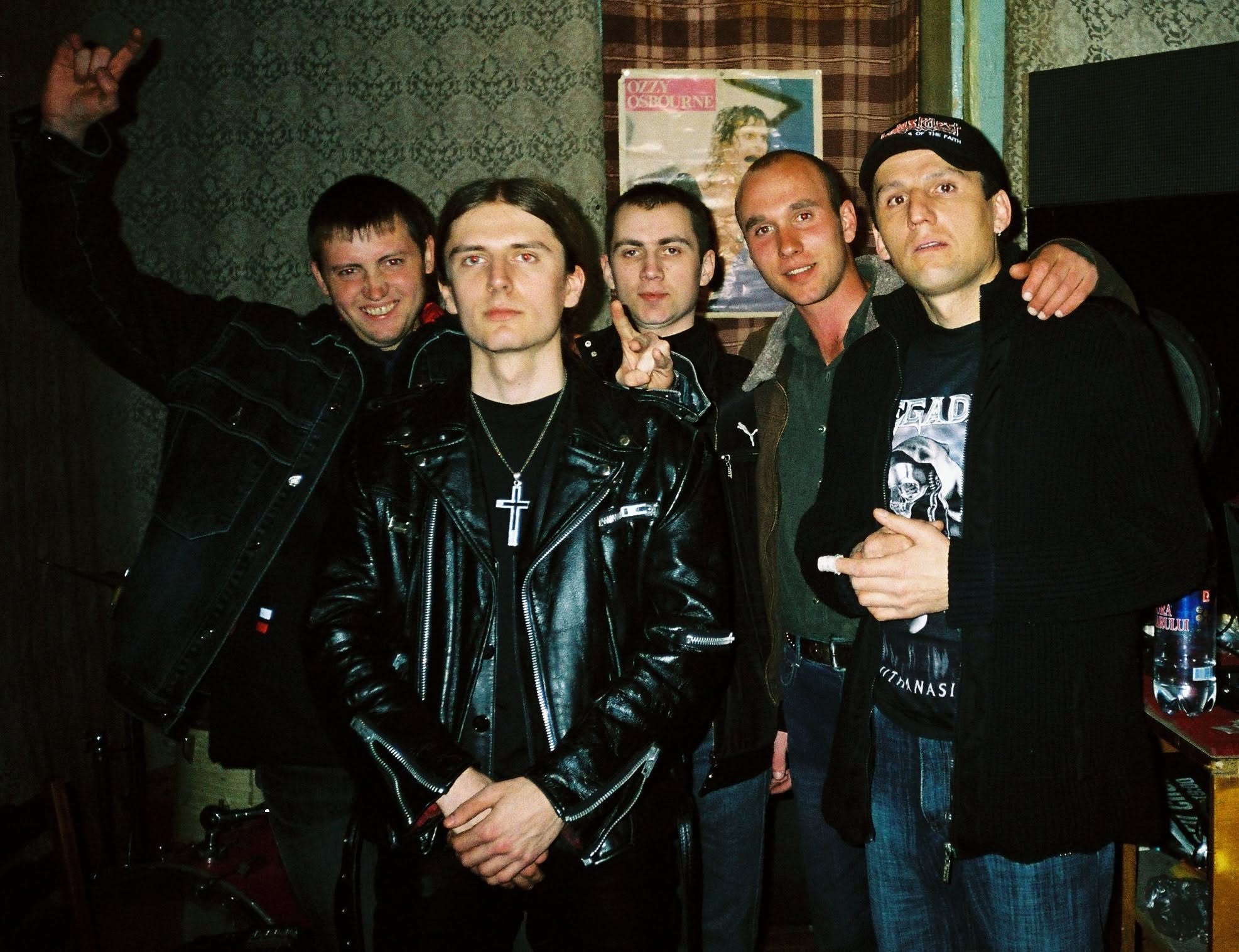 Group of five young men standing indoors, some smiling, one with arms raised, wearing casual clothing including leather jacket and baseball cap.