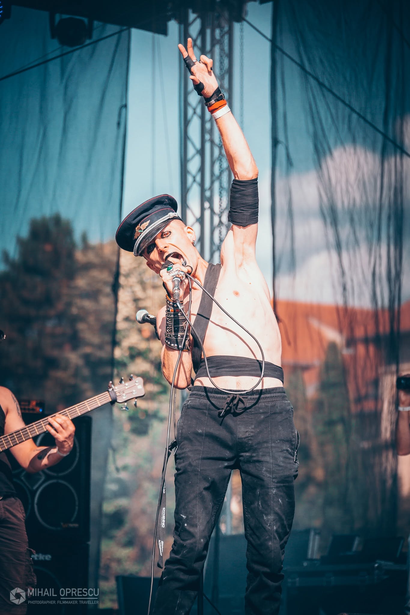 Male singer with glasses and a hat performs on stage, holding a microphone and raising one arm, with a guitarist nearby.