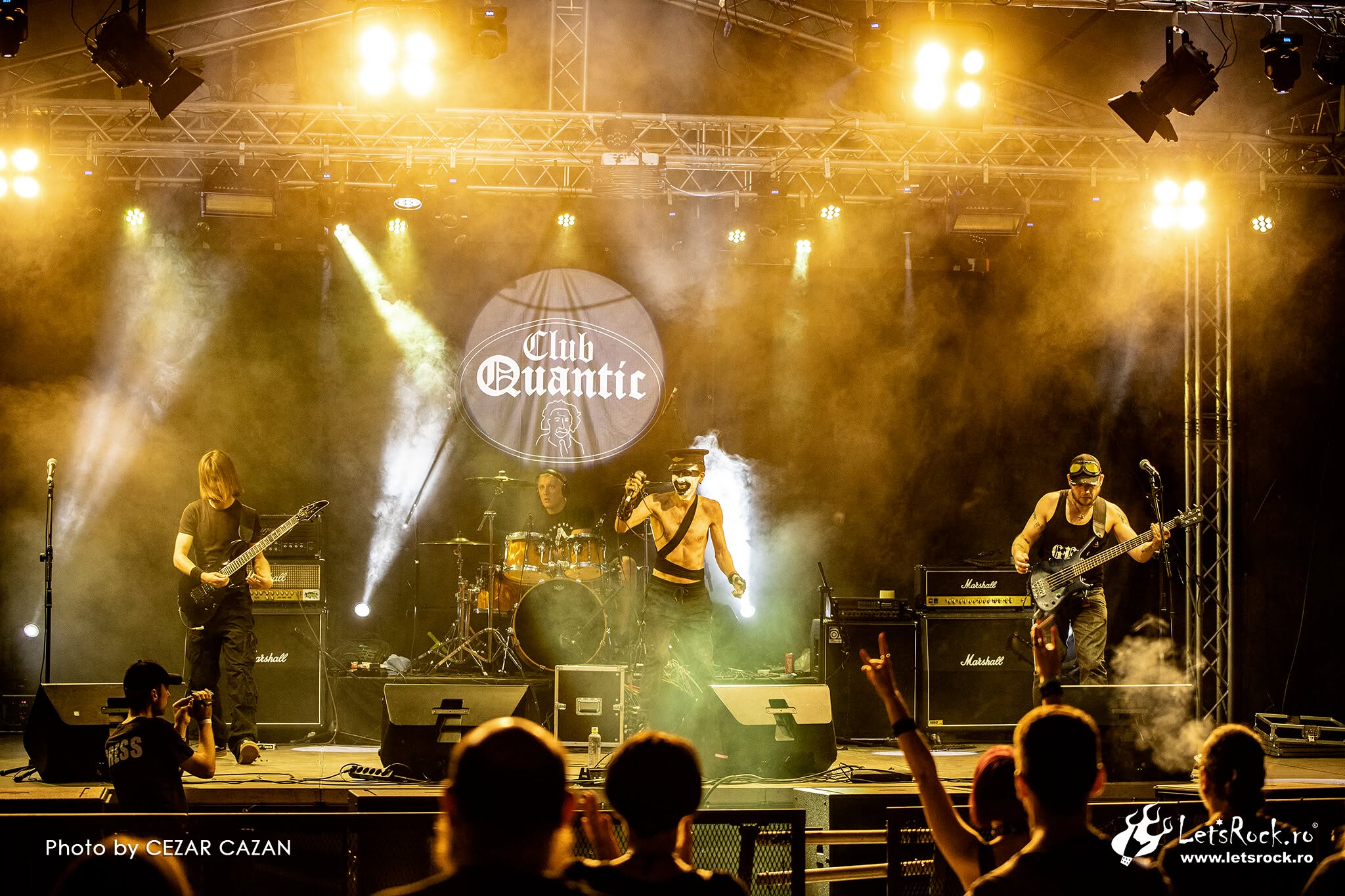 Band performs on stage with bright lights and fog, audience members watching and raising hands, club Quantic logo in background.