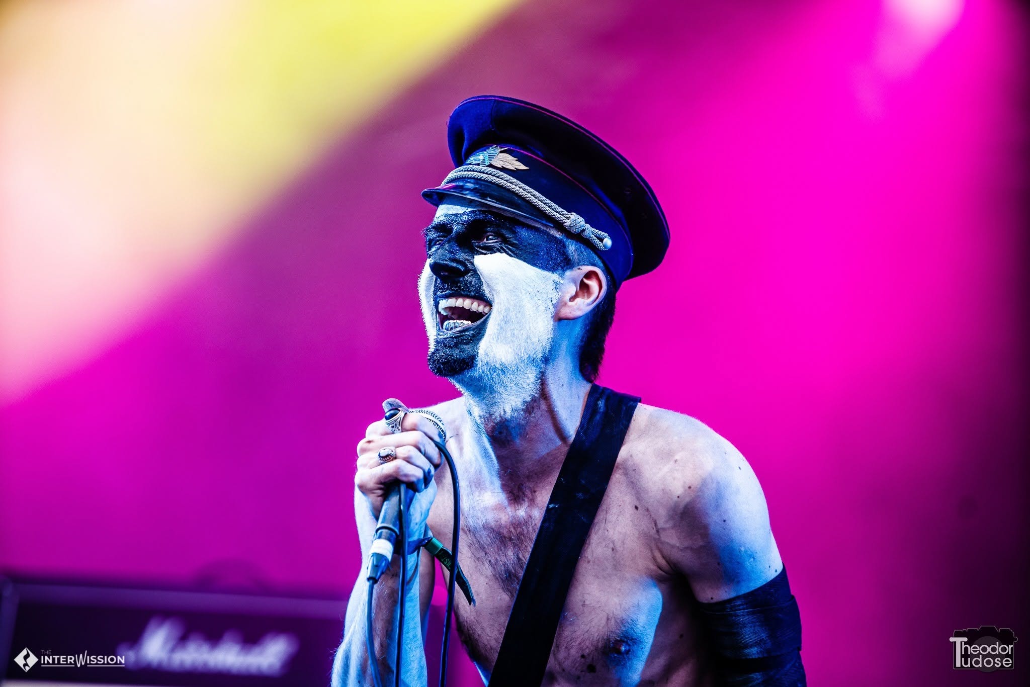 Person singing into a microphone on stage, wearing a hat and sunglasses, with colorful pink and yellow background.