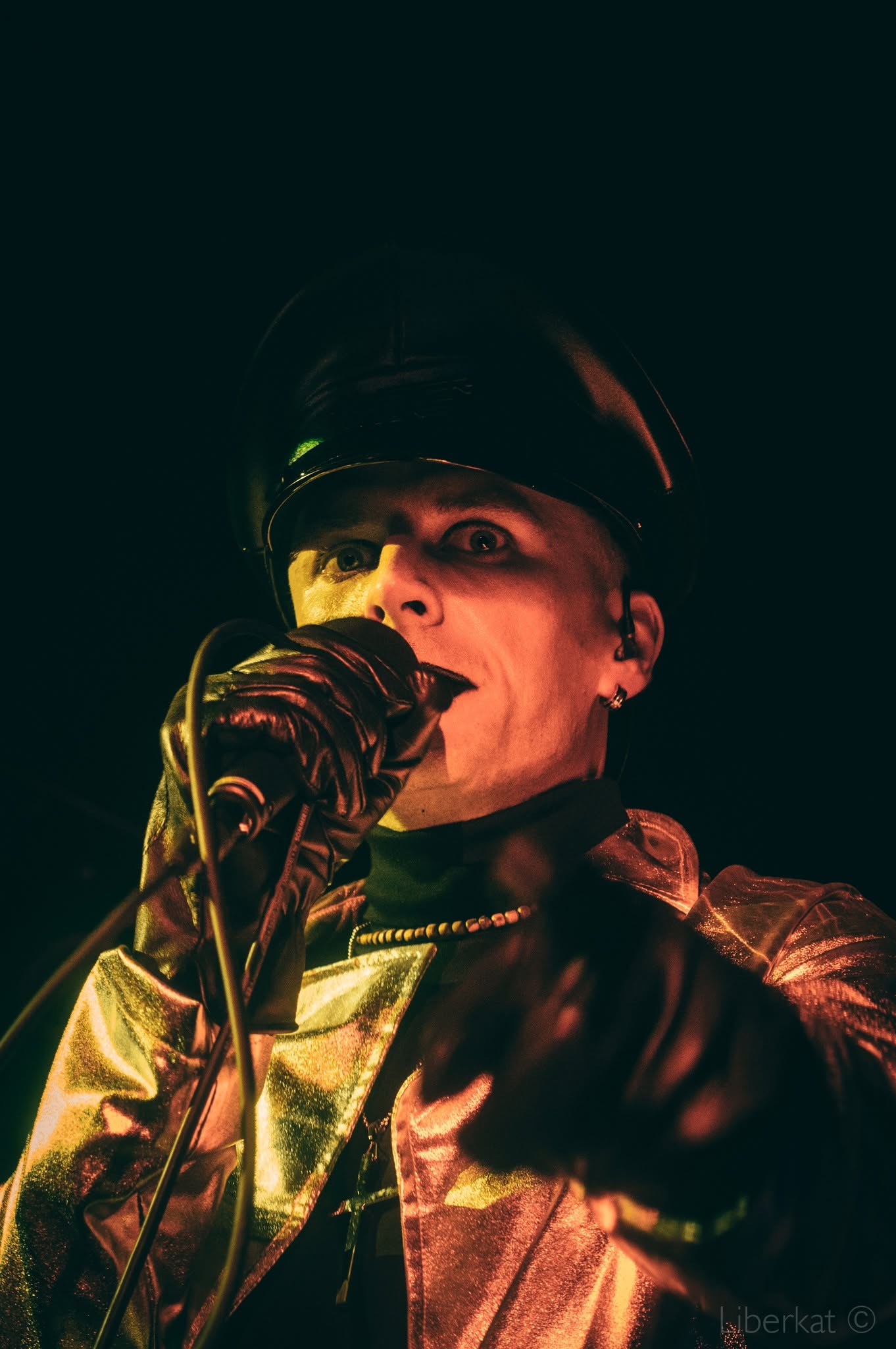Person wearing a hat and leather jacket singing into a microphone on stage, with dark background.
