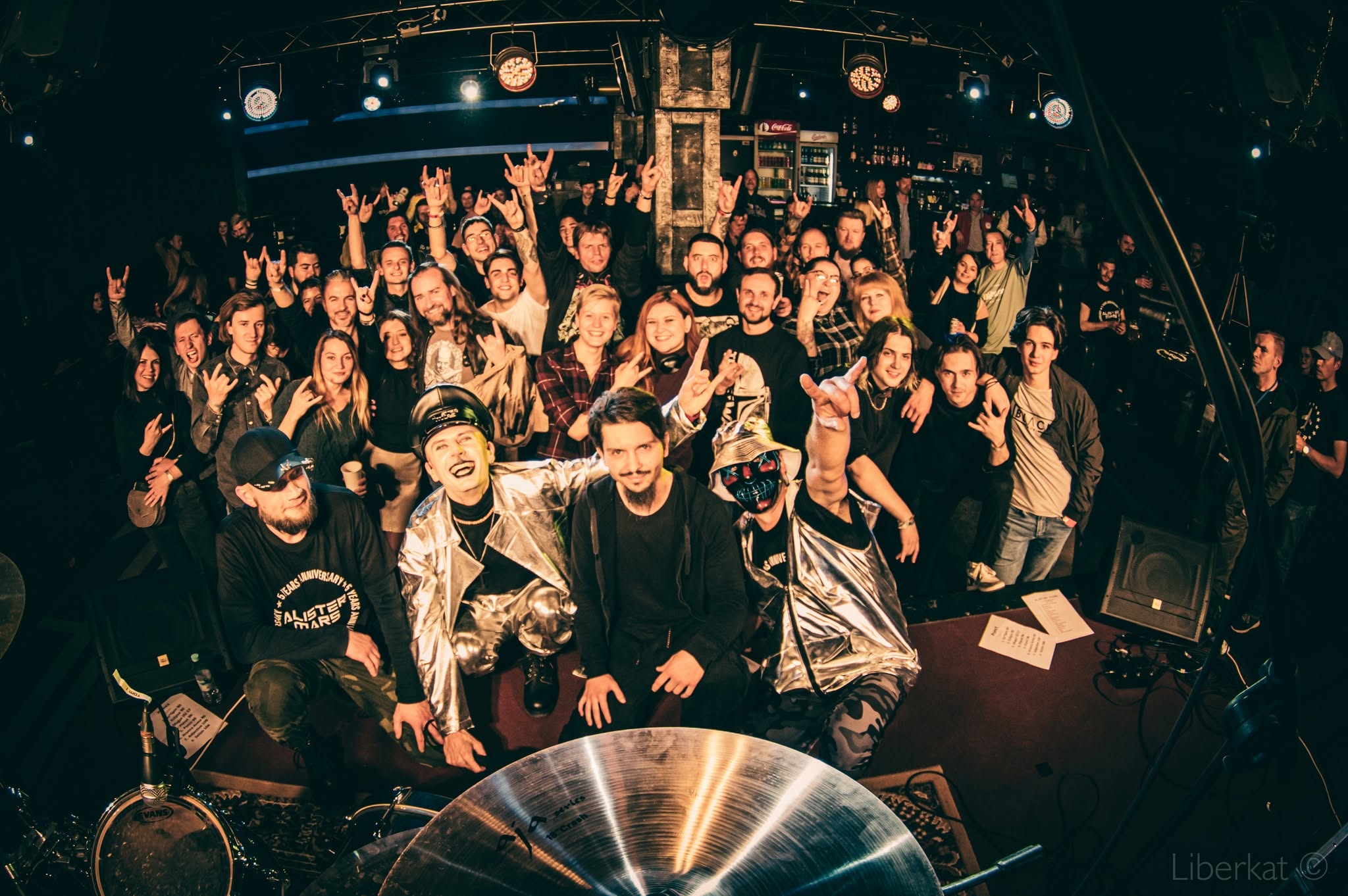 Crowd of people at a concert or event, some raising hands, in a dark venue with stage lighting.
