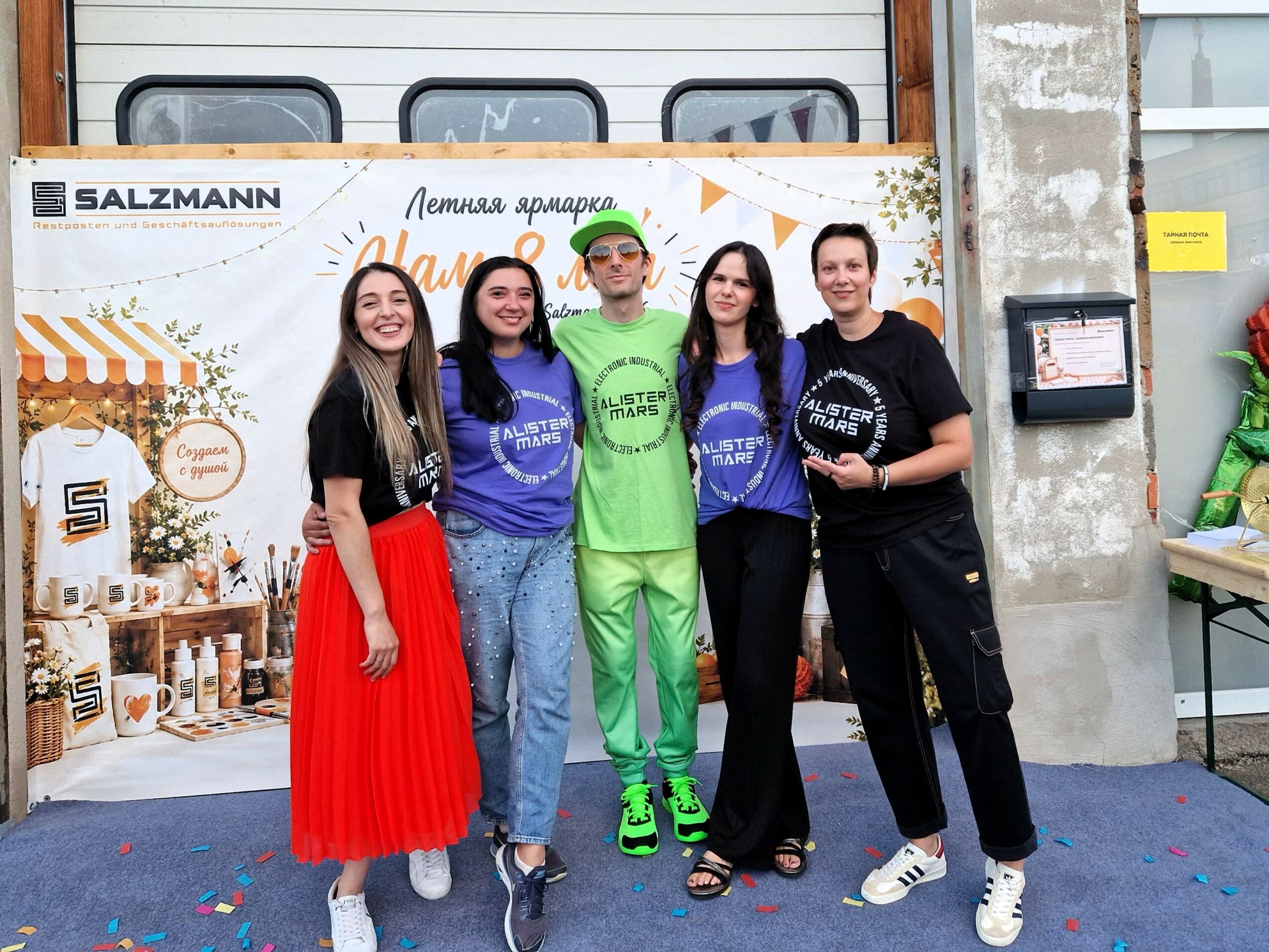 Five people standing together in front of a decorated backdrop, smiling, with a warehouse background.
