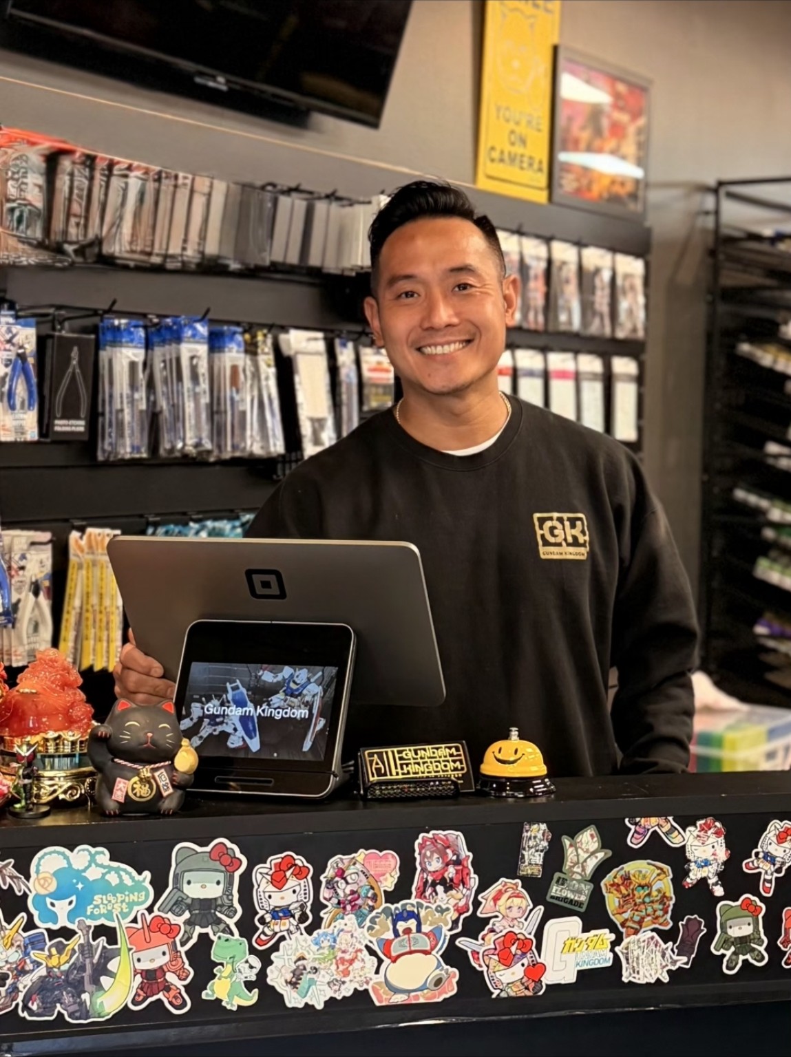 Smiling person standing behind a counter with stickers and a tablet, shelves with DVDs or games in background.