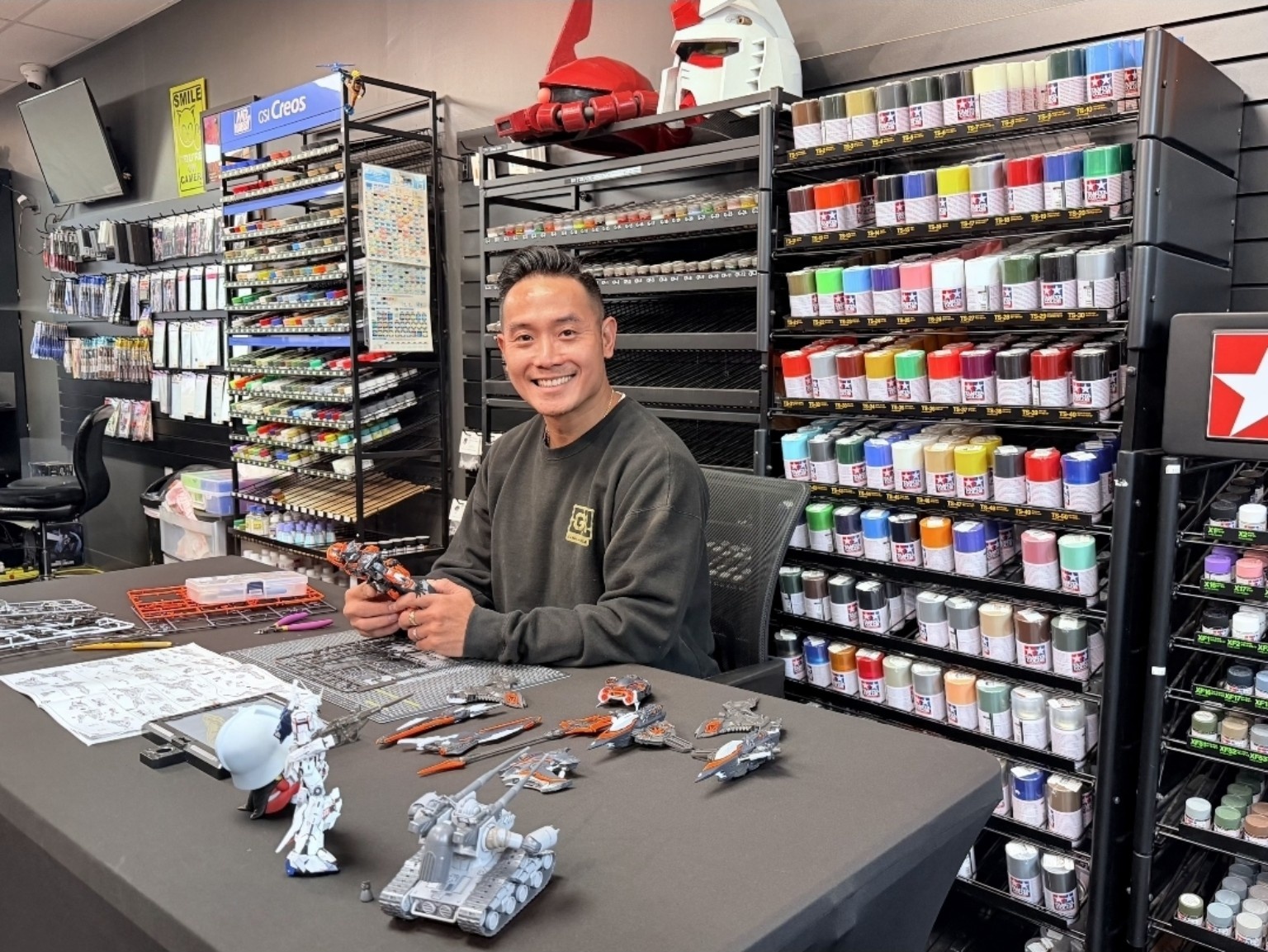 Smiling person sitting at a table with model parts and tools, surrounded by shelves of paints and supplies in a store.