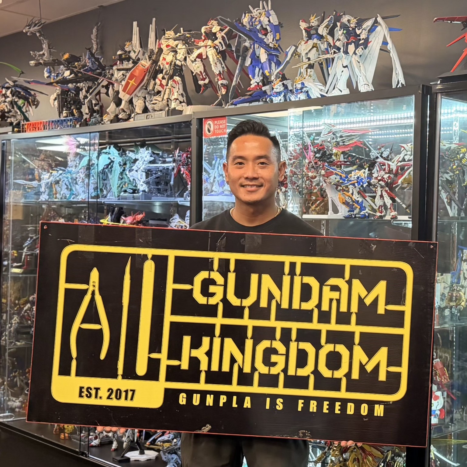 Smiling person holding a large black and yellow sign with text about Gundam and a display of Gundam models in the background.