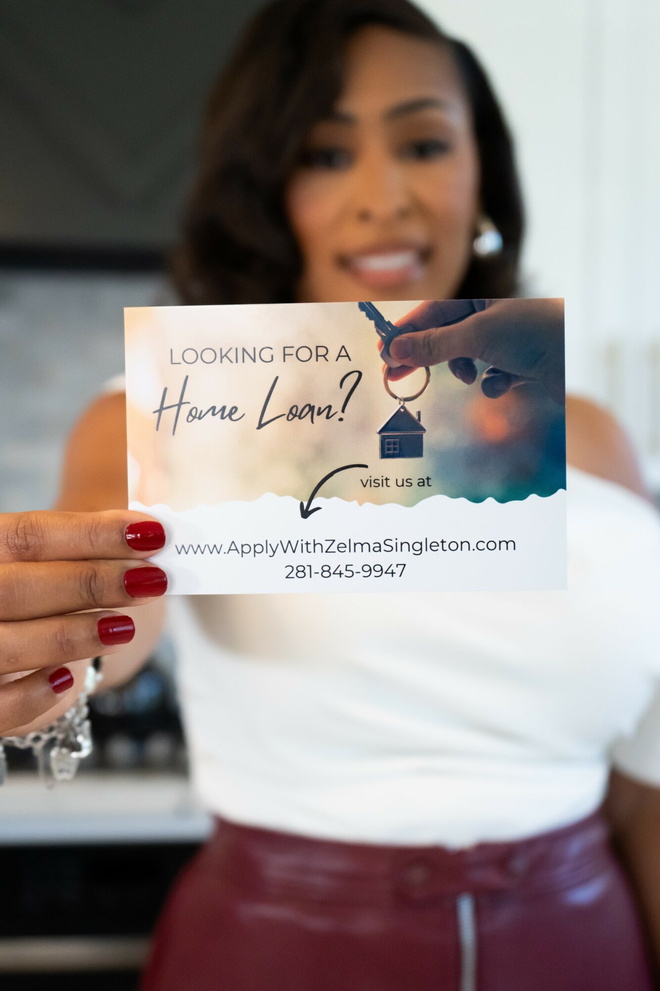 Person holding a business card with a house drawing and text about home loan, in front of a blurred woman background.