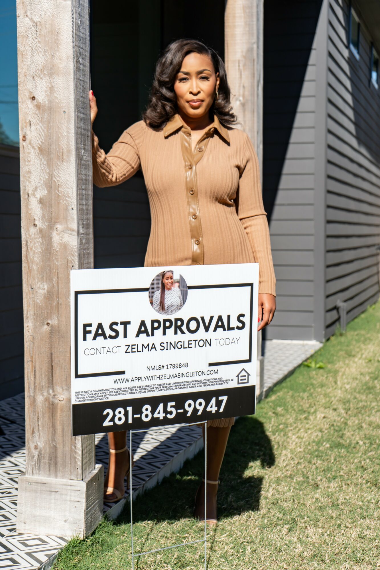 Woman standing outdoors holding a sign with contact information and a photo, next to a wooden post and modern building, sunny day.