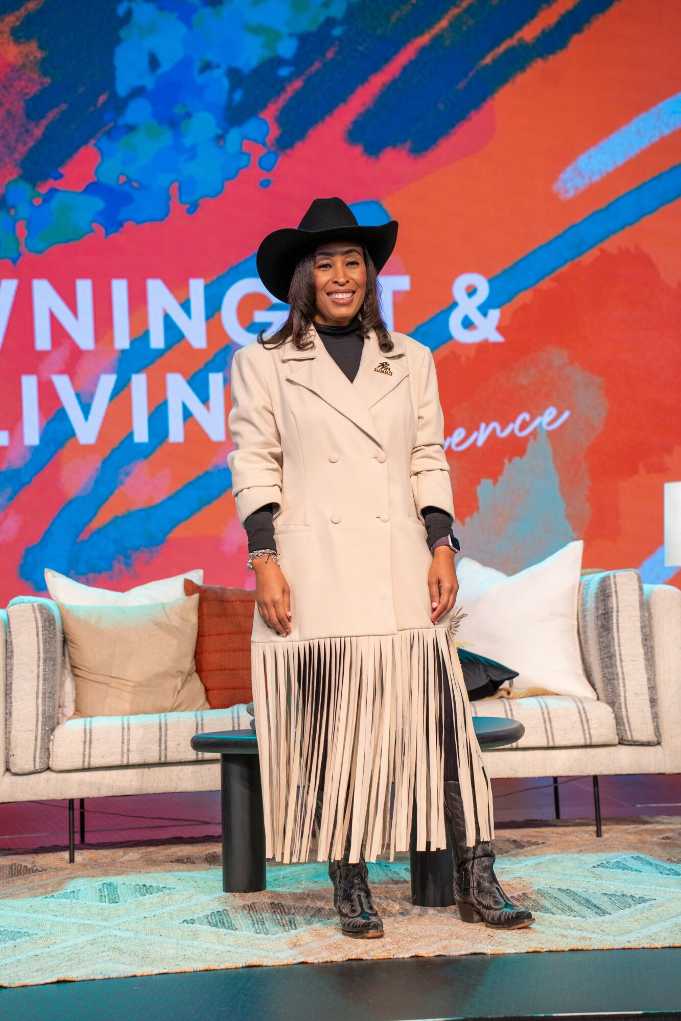 Woman in beige coat and black hat standing on stage with colorful background and sofa behind her.