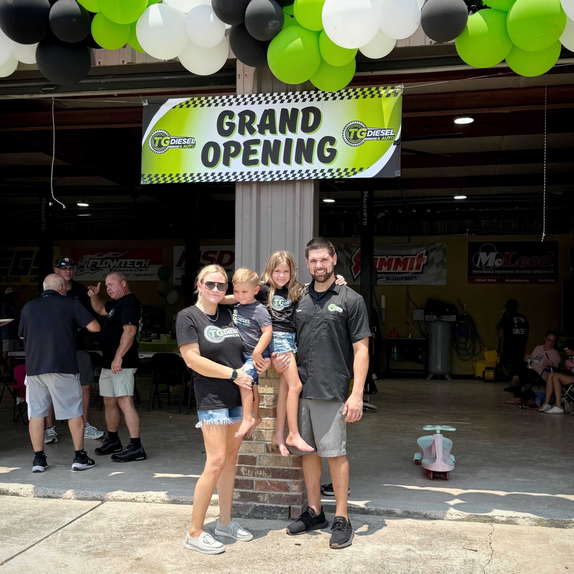 Group of four people, including a child, standing in front of a grand opening sign with balloons overhead.