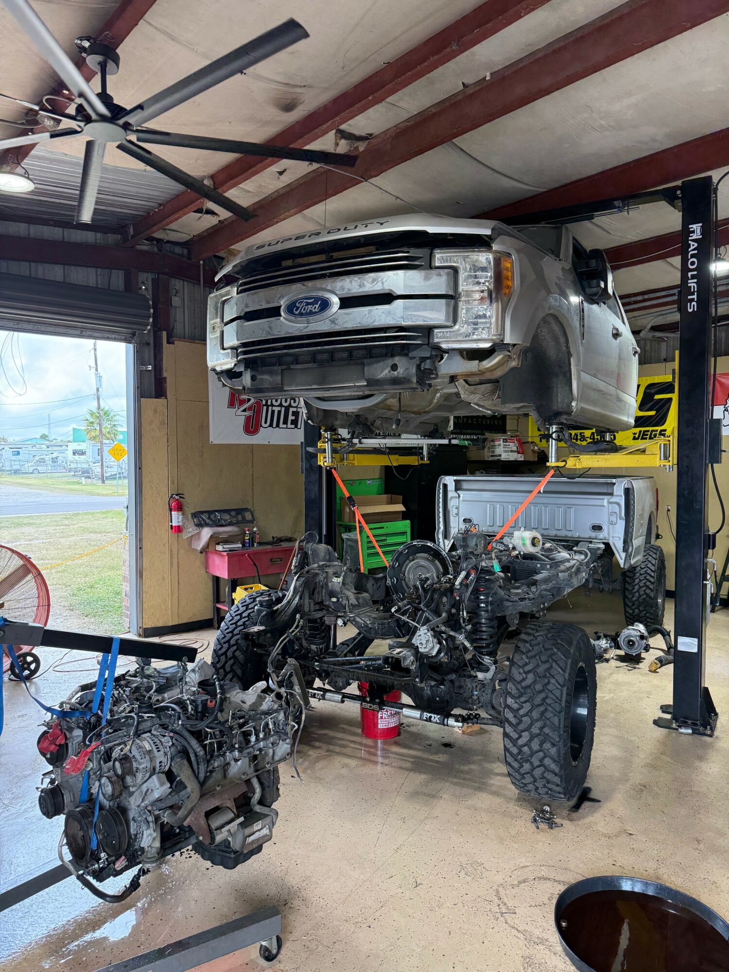 Car lifted on a hydraulic lift inside a garage, with engine parts on the floor, and a ceiling fan overhead.