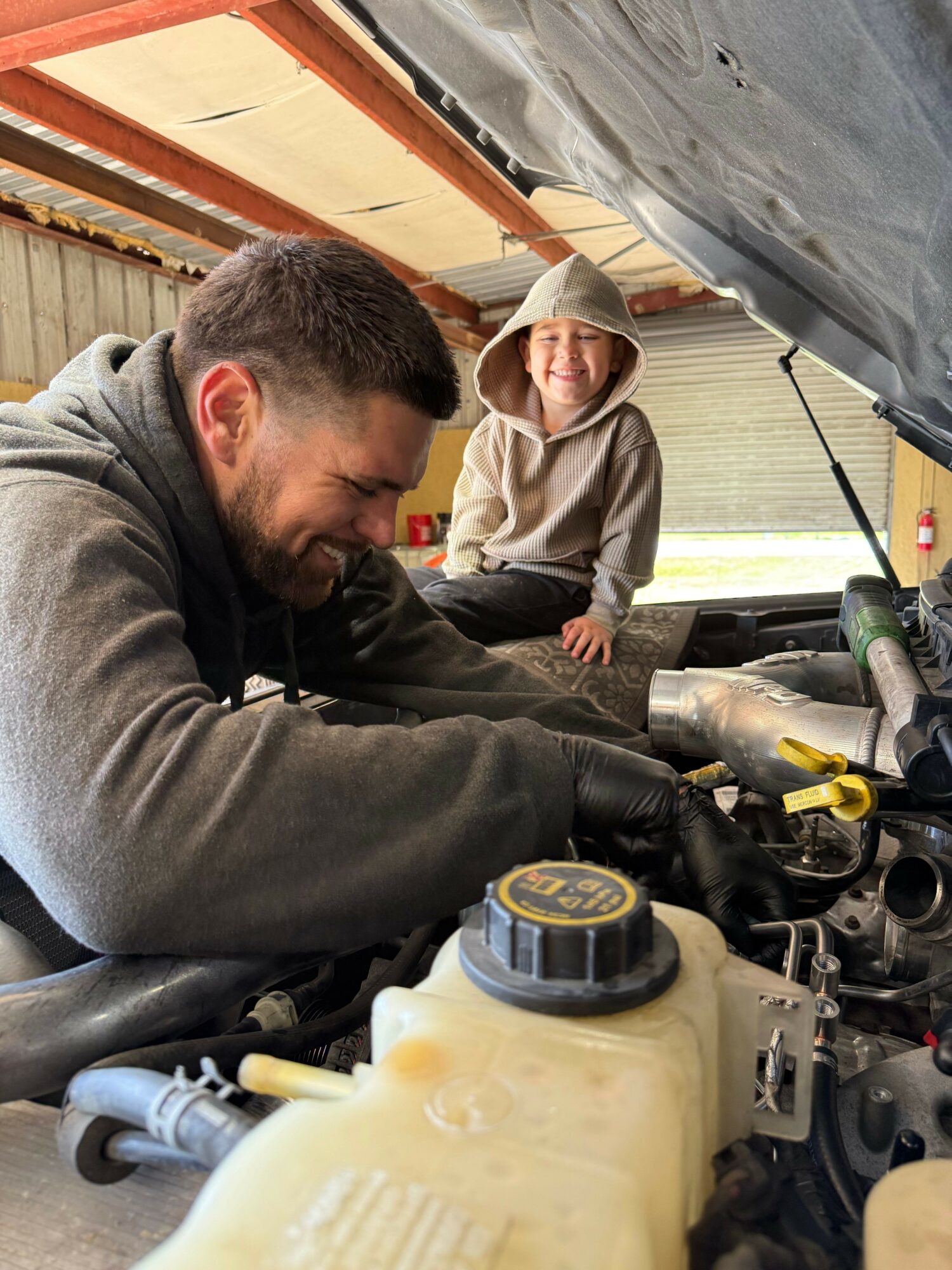 Man working on car engine with a child observing, engine components visible, indoors, focused on repair.