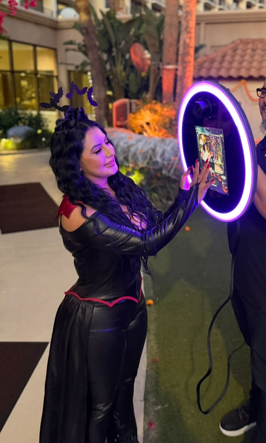 Woman with dark curly hair and black outfit interacts with a circular illuminated mirror at an indoor event.