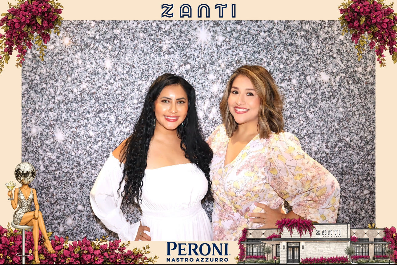 Two women standing in front of a glittery backdrop with flowers and a sign, smiling at the camera.
