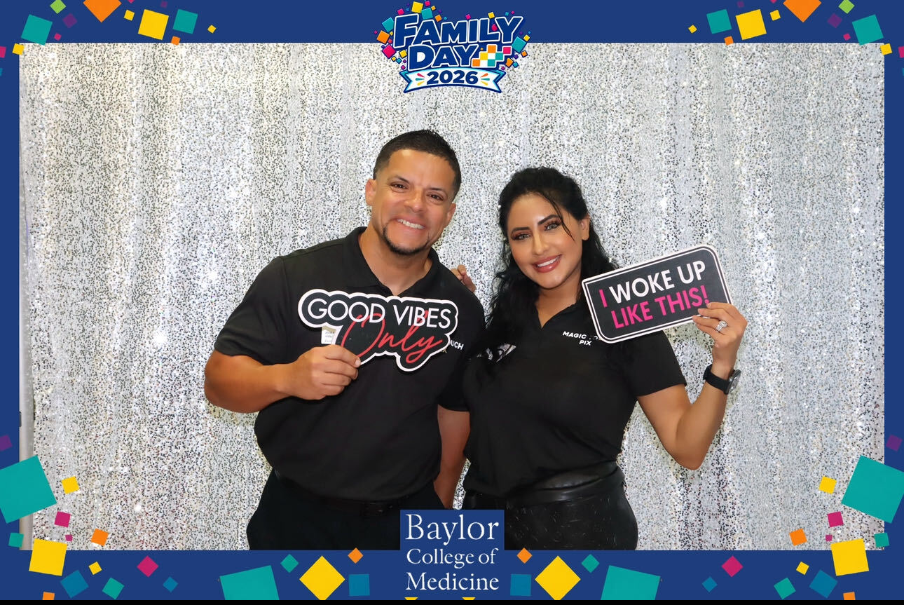 Two people smiling, holding signs, standing in front of a silver curtain at Family Day event, Baylor College of Medicine.