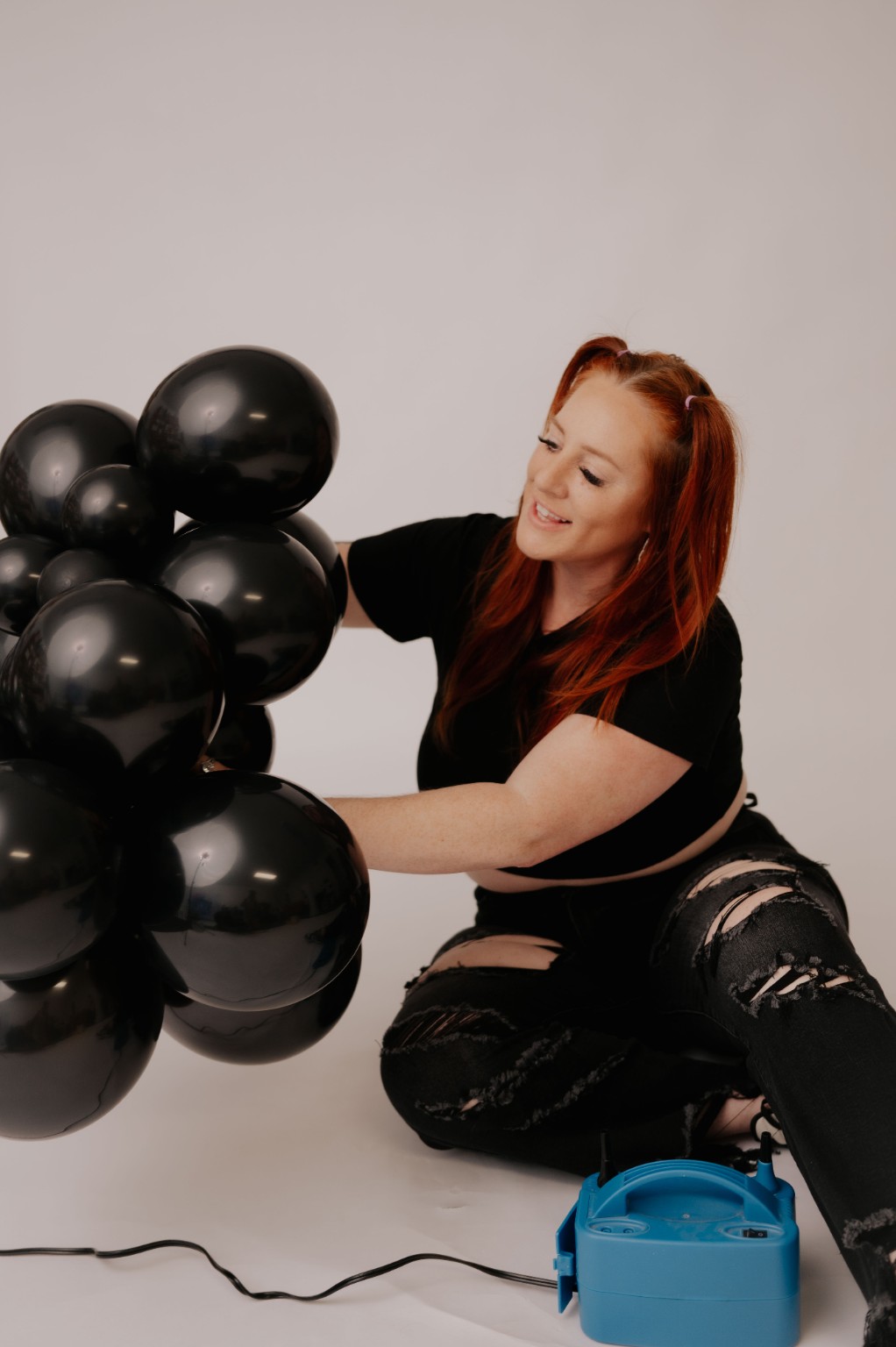 Woman with red hair sitting on the floor, holding black balloons, against a plain light background.