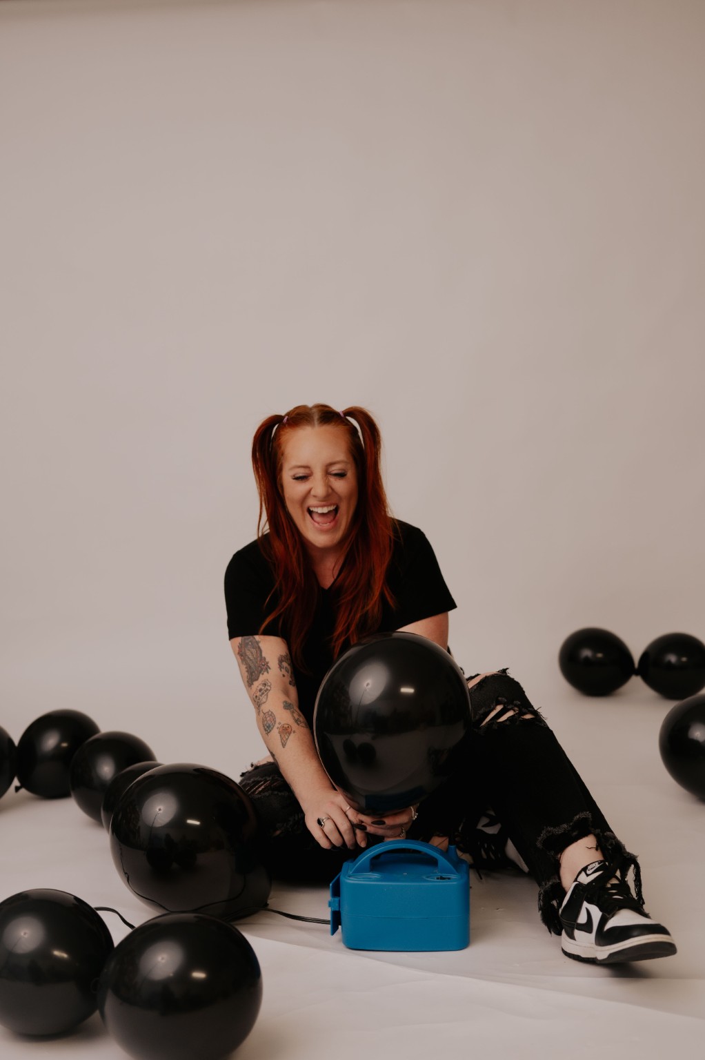 Person with red hair and tattoos sitting on the floor, smiling, surrounded by black balloons, with a blue object nearby.