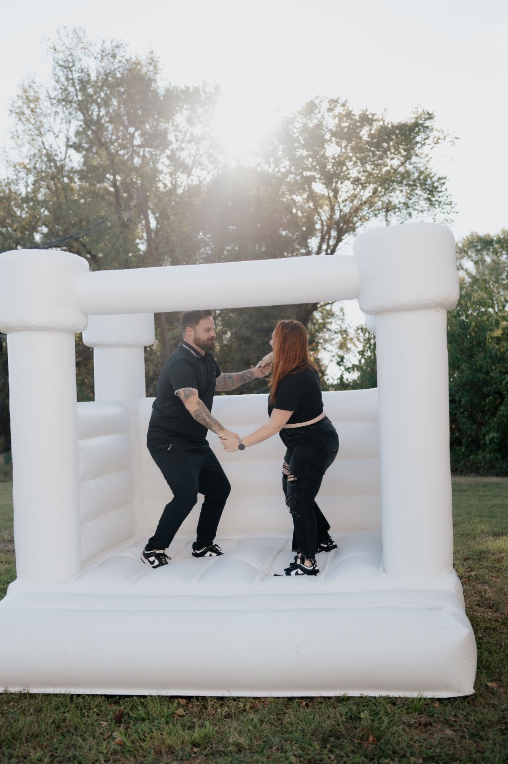 Two people holding hands and dancing on an inflatable bounce house outdoors, with trees and sunlight in the background.
