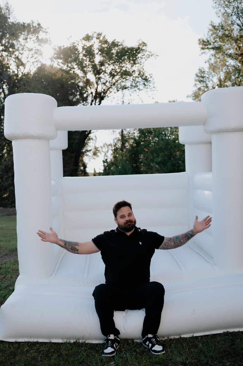 Man with tattoos kneeling in front of a large white inflatable bounce house outdoors, trees in background.