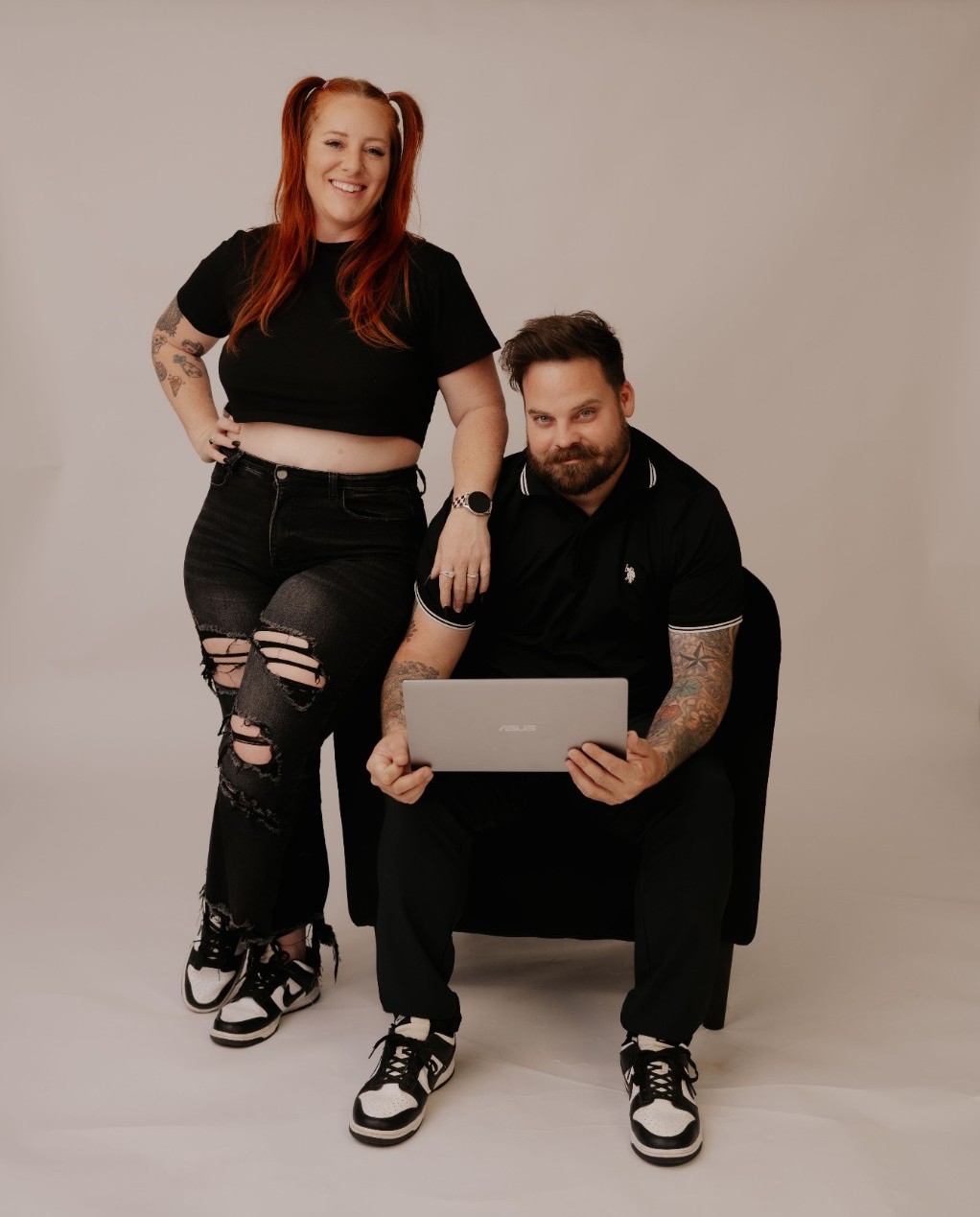 A smiling woman with red hair and a man with a beard sit together, woman standing and man sitting with a laptop, against a plain background.