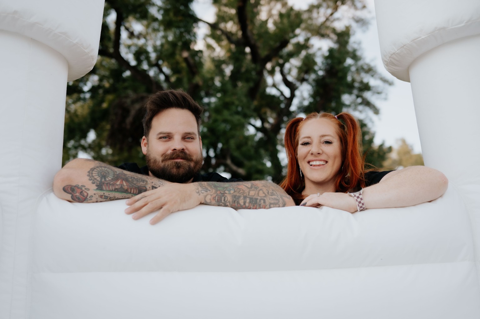 A man and woman leaning on a white inflatable structure outdoors, smiling, with trees in the background.