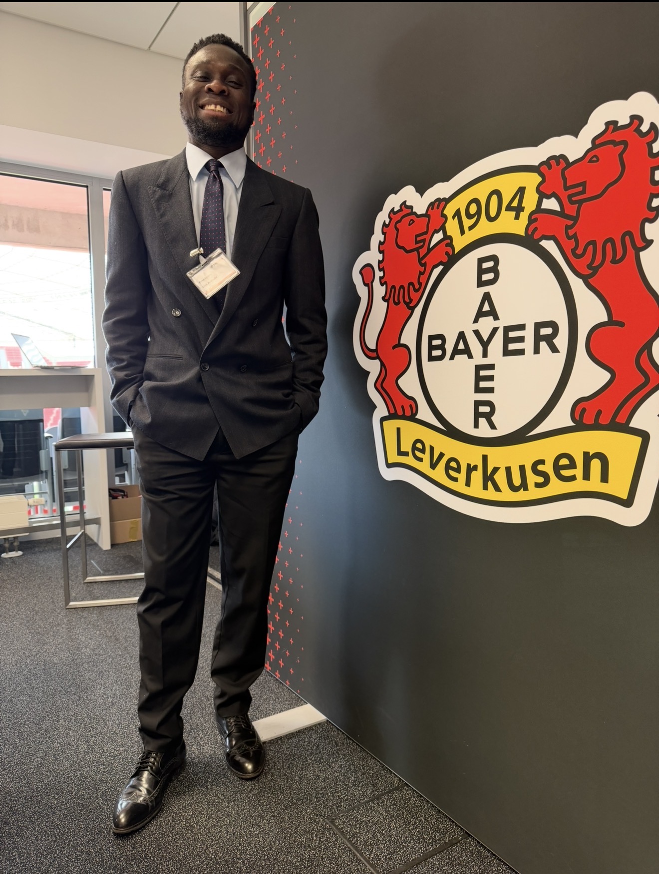 Man in suit standing next to Bayer Leverkusen logo on wall, smiling, in an indoor setting.