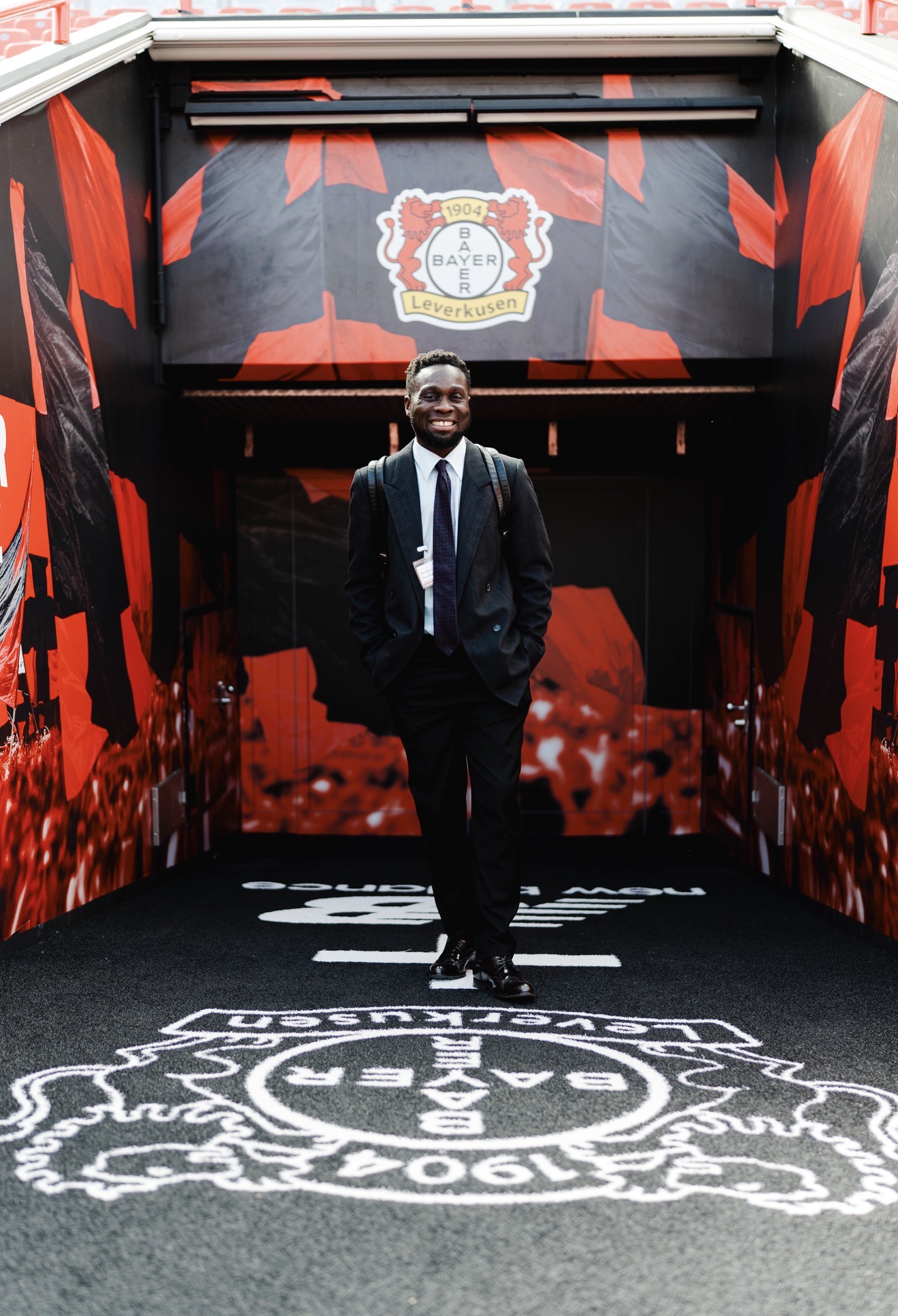 Man in suit walking through a decorated tunnel with red and black patterns and a logo overhead, smiling confidently.