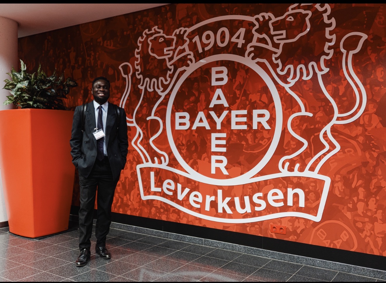 Man in suit standing next to a large Bayer Leverkusen logo on a red wall.
