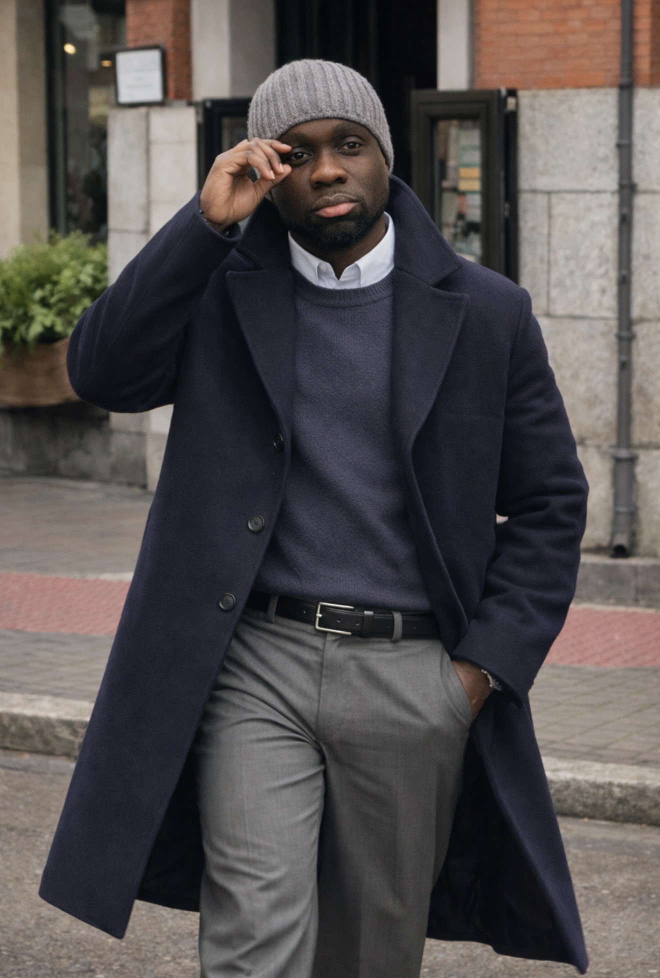 Man in coat and gray beanie adjusting glasses on street, with brick building and sidewalk in background.