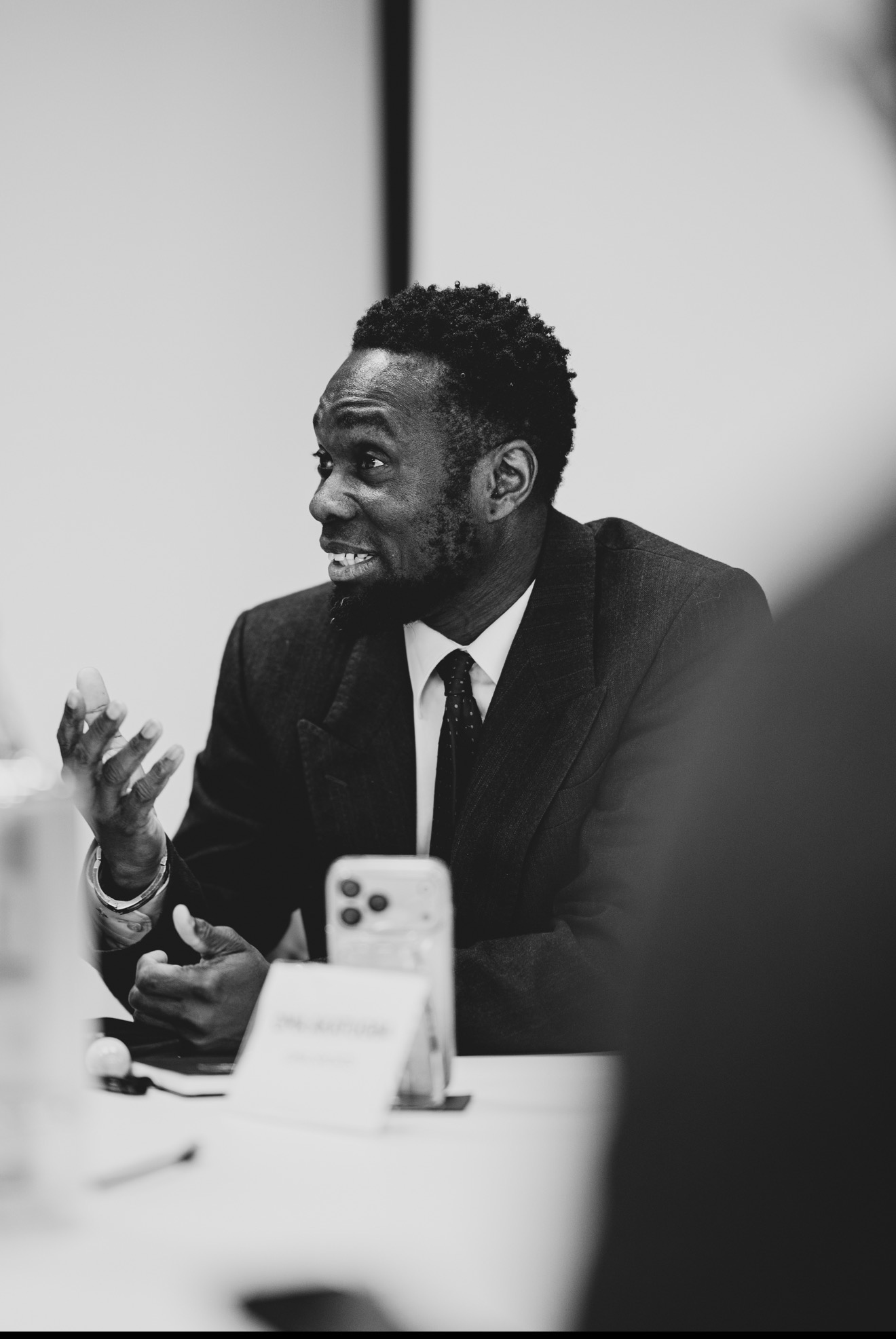 Man in suit speaking and gesturing during a meeting, with a smartphone and nameplate on table, black and white image.