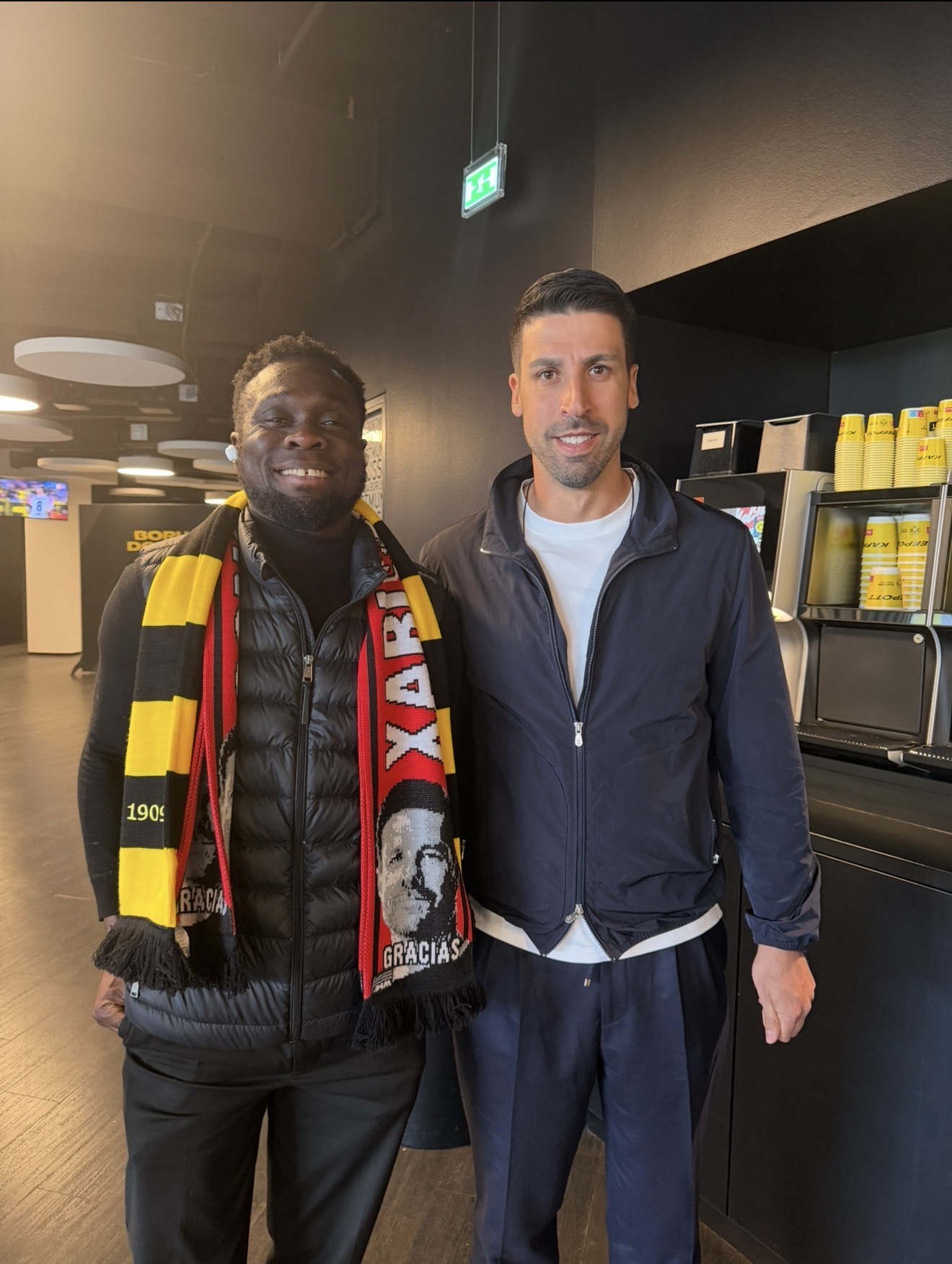 Two men standing side by side indoors, one wearing a black jacket and scarf, the other in a navy jacket, smiling.