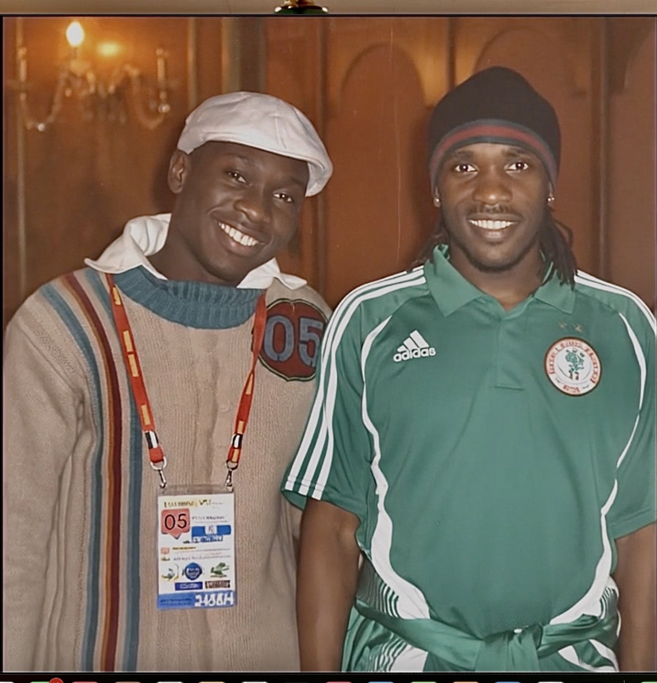 Two smiling men standing indoors, one wearing a white head covering and colorful sweater, the other in a green sports jersey.