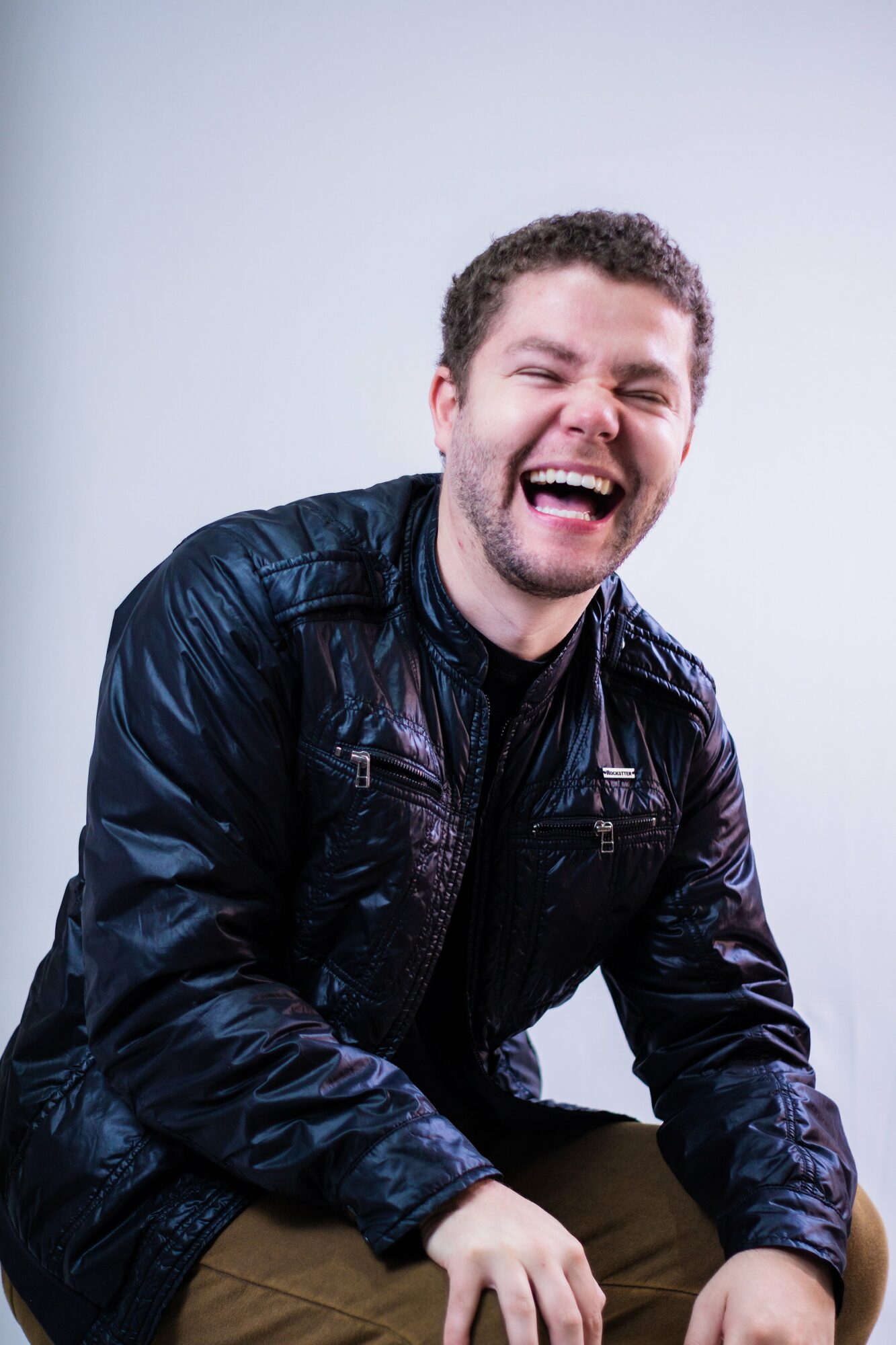 Young man with curly hair laughing, wearing a dark jacket, against a plain light background.