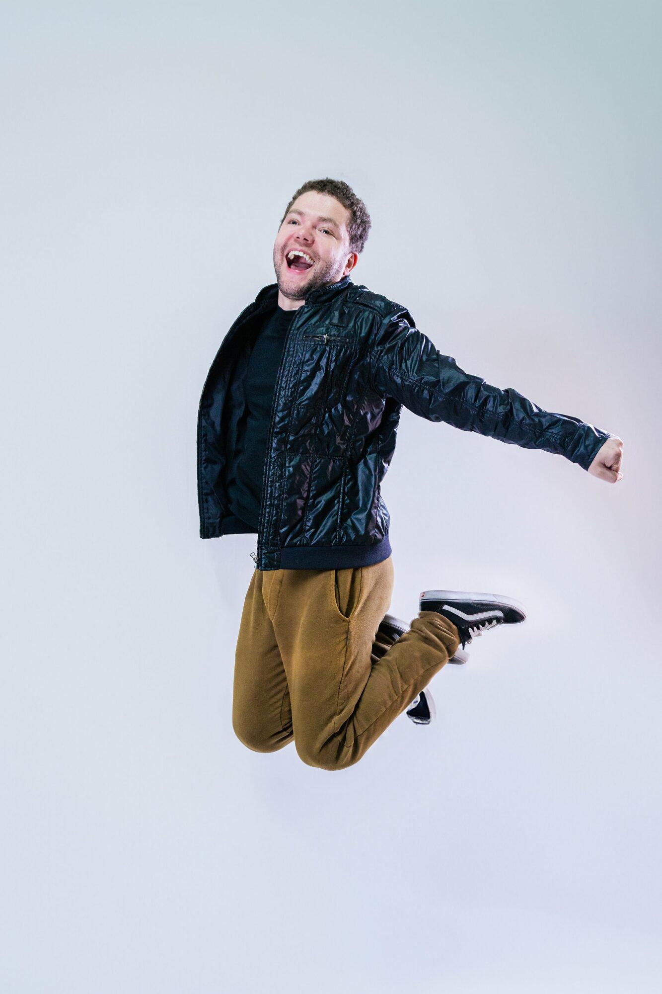 Person mid-air with arms outstretched, smiling, wearing a black jacket, brown pants, and black shoes, against a plain background.