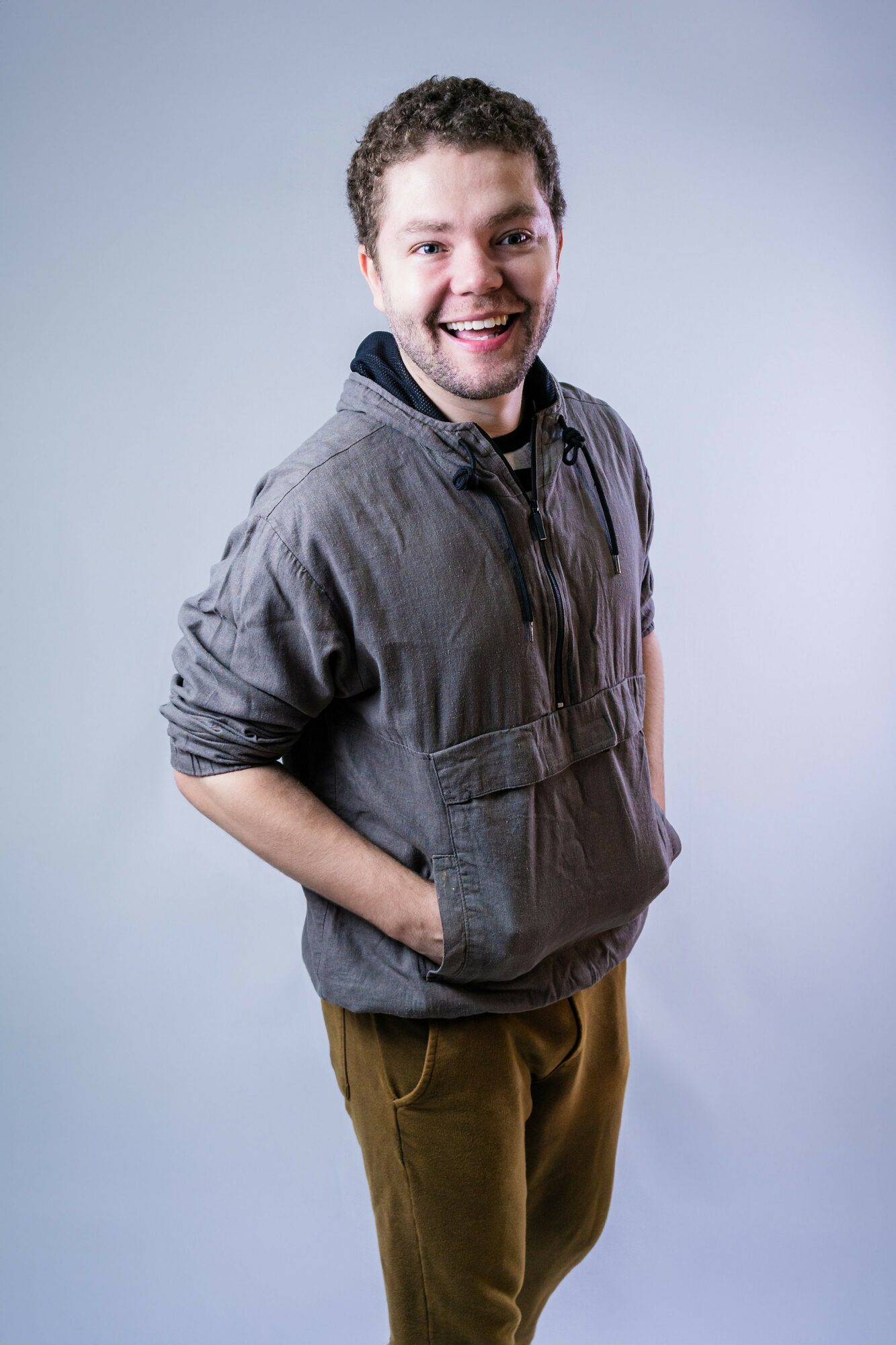 Young man with curly hair smiling, wearing a gray jacket and brown pants, standing with hands in pockets against a light background.