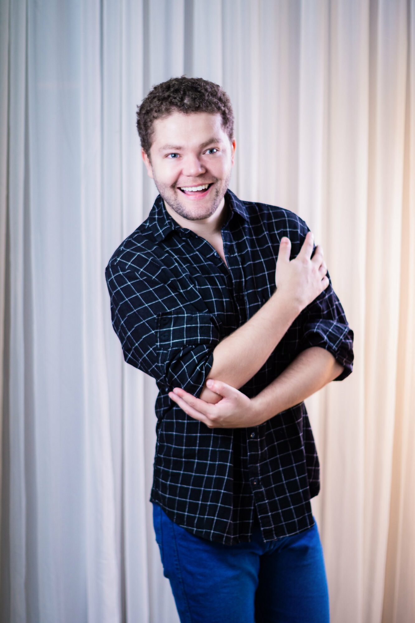 Young man with curly hair smiling, wearing a dark checkered shirt and blue pants, standing in front of curtains.