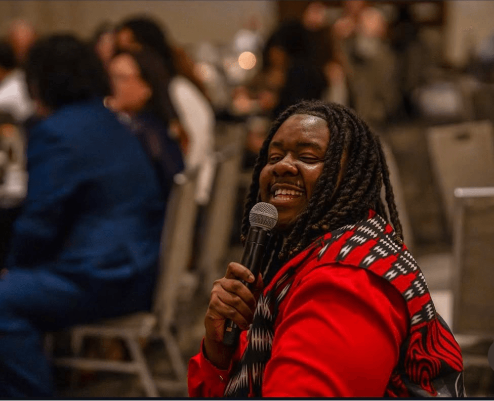 Person with long braided hair smiling and holding a microphone, wearing a red jacket, in a crowded indoor setting.