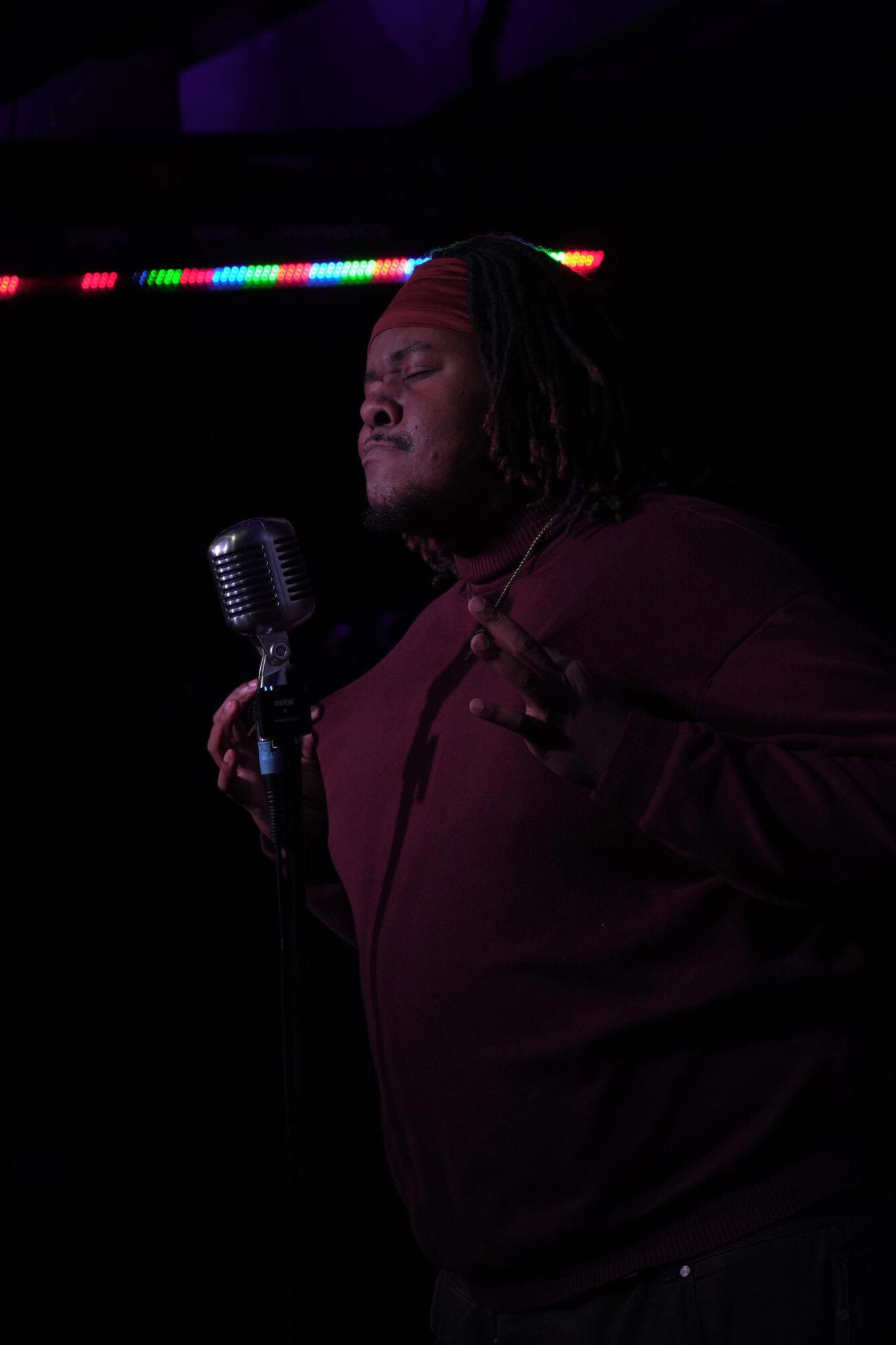 Person singing into a vintage microphone with closed eyes, dark background, colorful lights above, wearing a maroon top.