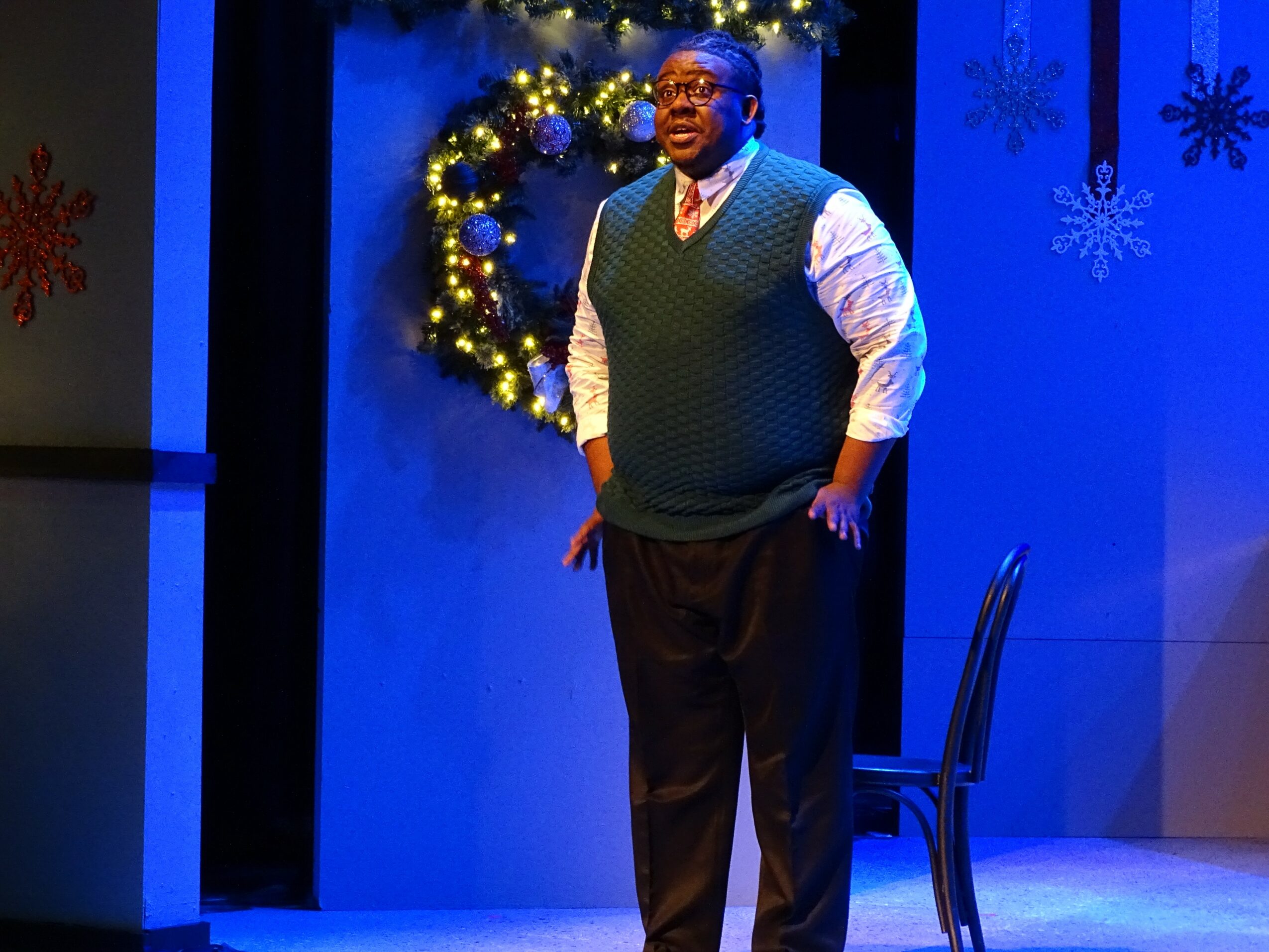 Man standing on stage with holiday decorations, wearing glasses, a vest, and dress pants.