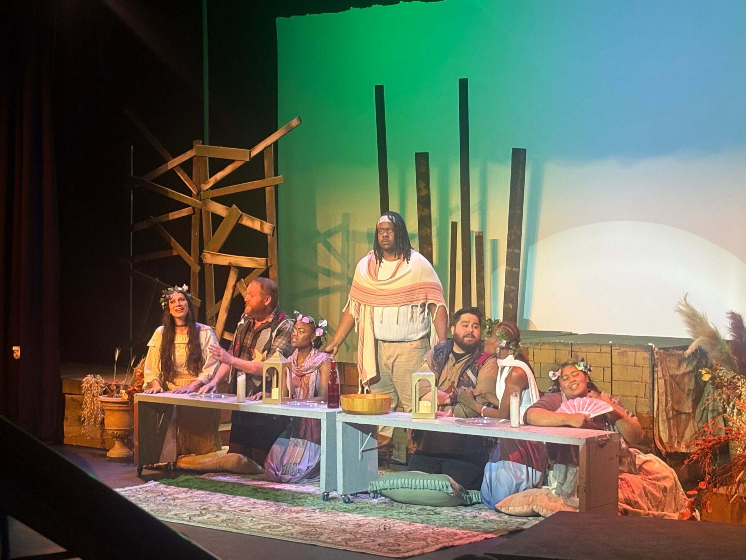 Group of people performing on stage with wooden props and a colorful backdrop, some seated at a table, others standing.
