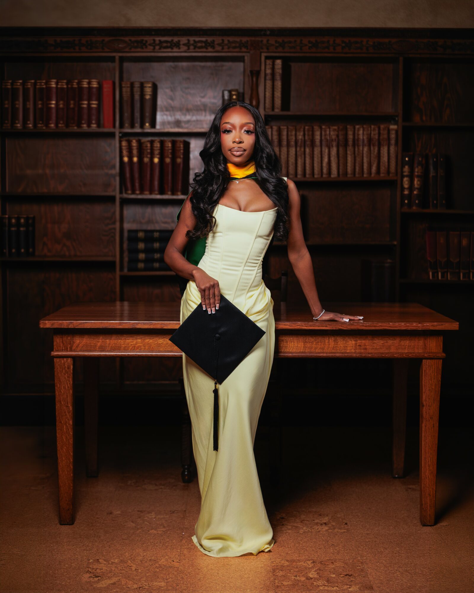 Woman in a cream-colored dress holding a black clutch, standing in front of a wooden table and bookshelf.